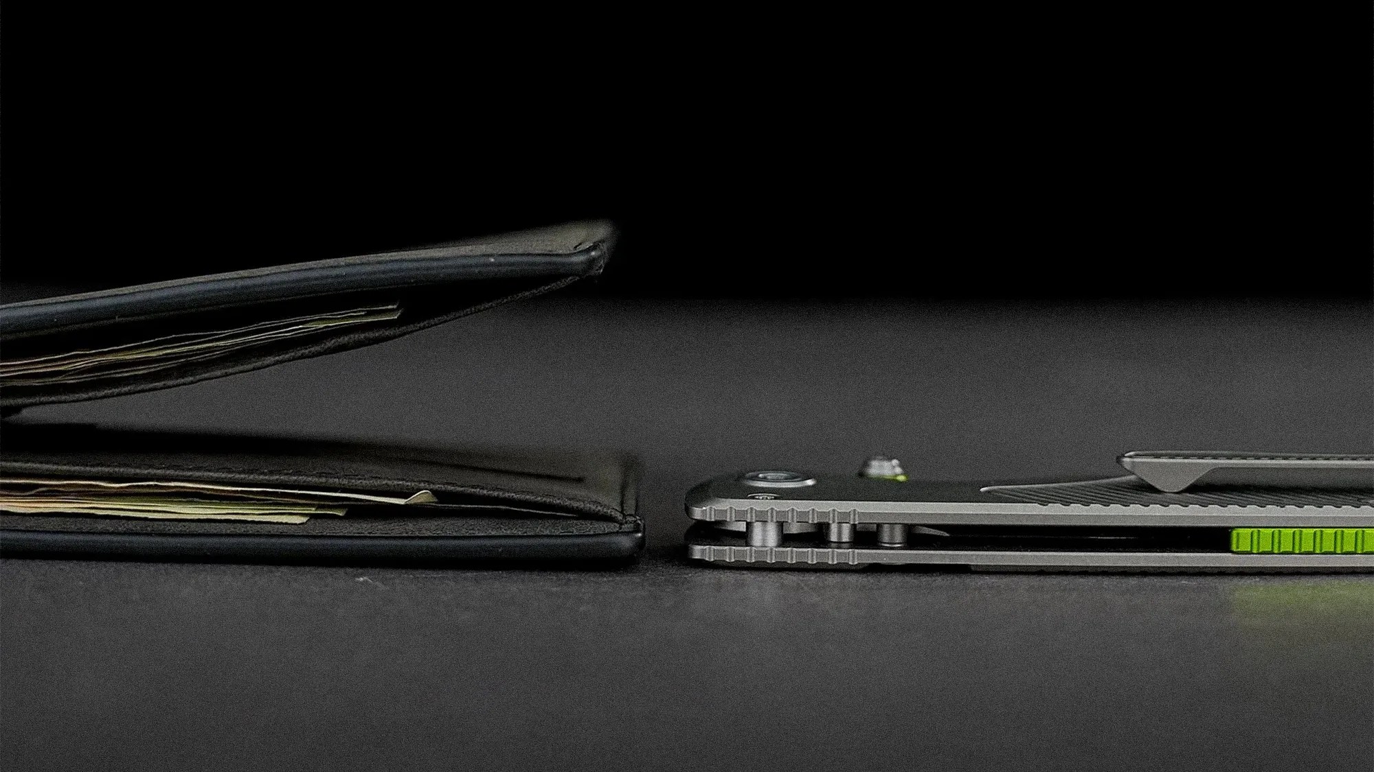 Side view of a thick black leather wallet filled with cash next to a slim closed folding knife with a metal clip and green accents on a dark surface.