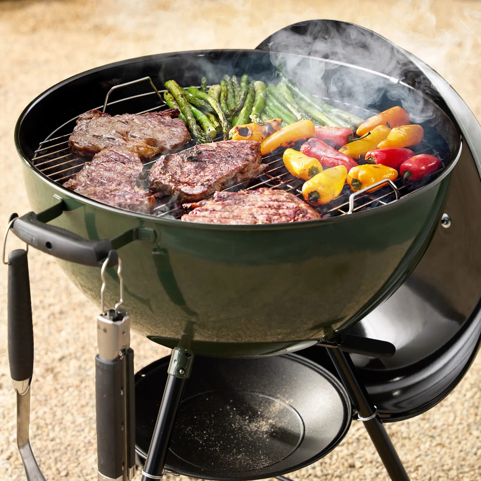 Green charcoal kettle grill with steaks, asparagus, and colorful mini bell peppers cooking on the grate with smoke rising.
