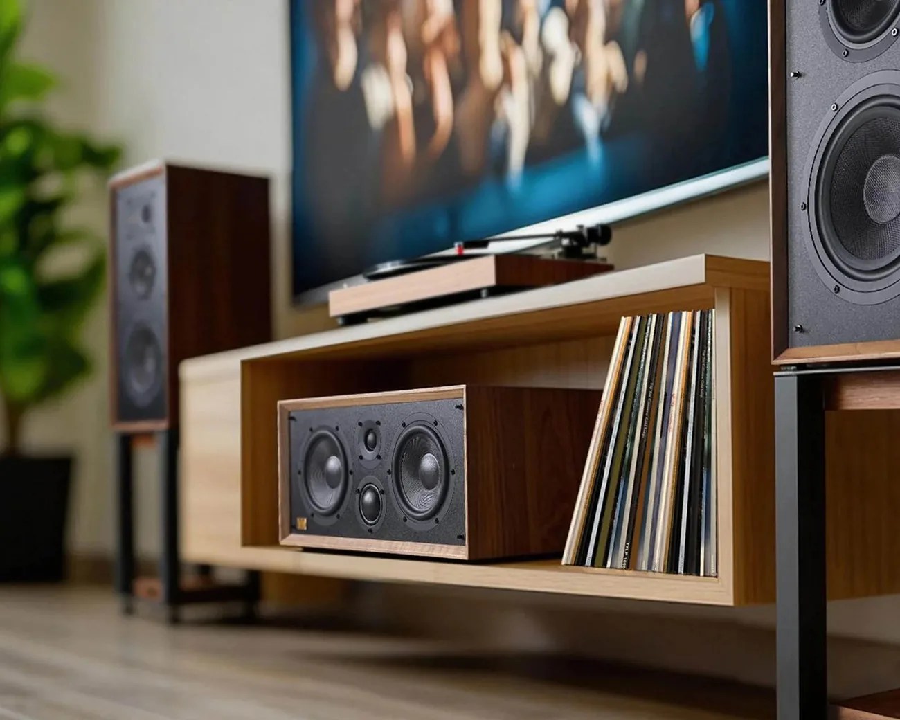 Wooden center speaker and vinyl records on a light wood media console with floor-standing speakers and a turntable in the background.