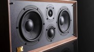 Rectangular speaker with two large black woofers, two smaller tweeters, and a wooden frame on a reflective black surface.