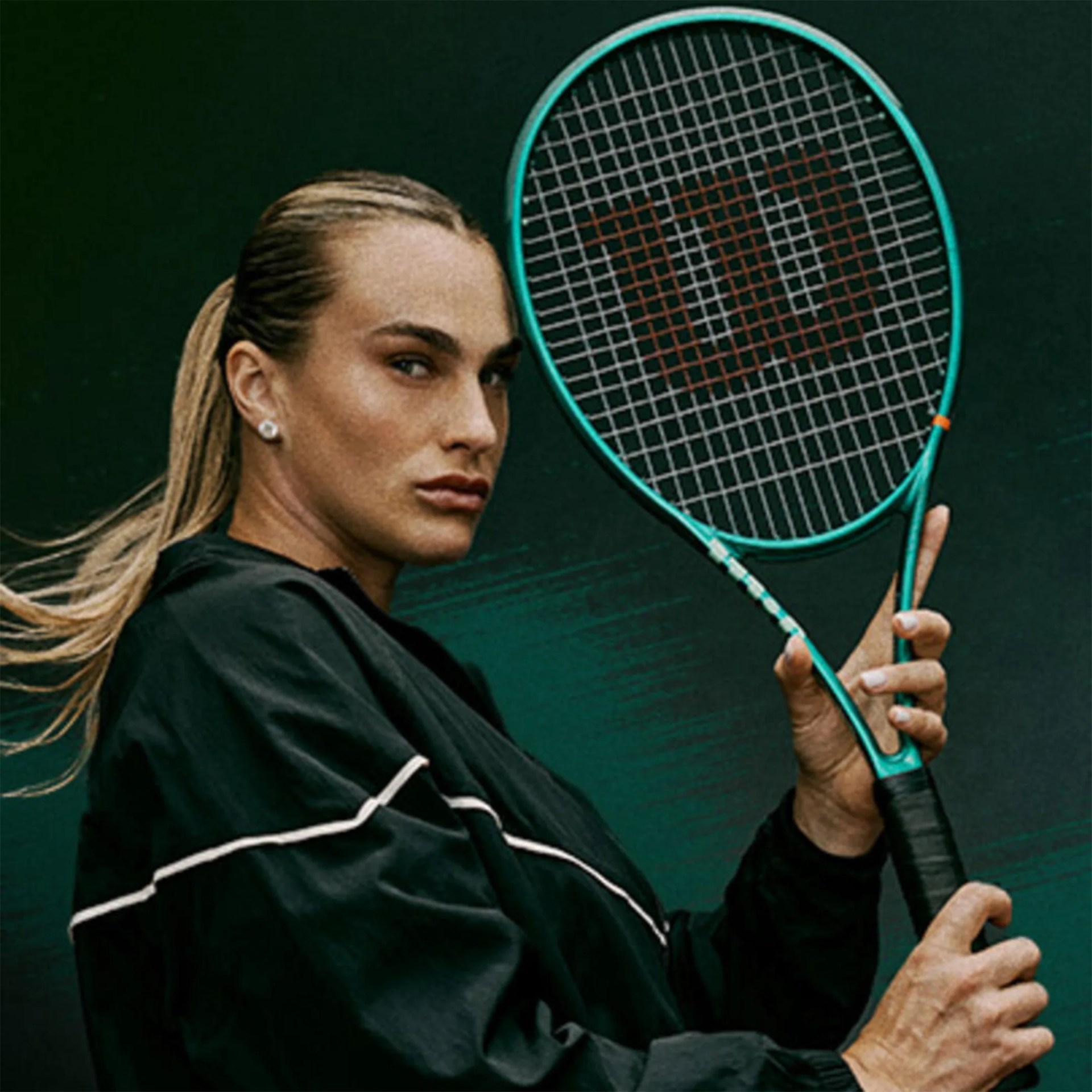 Person with long hair tied back wearing a black jacket holding a teal Wilson tennis racket.