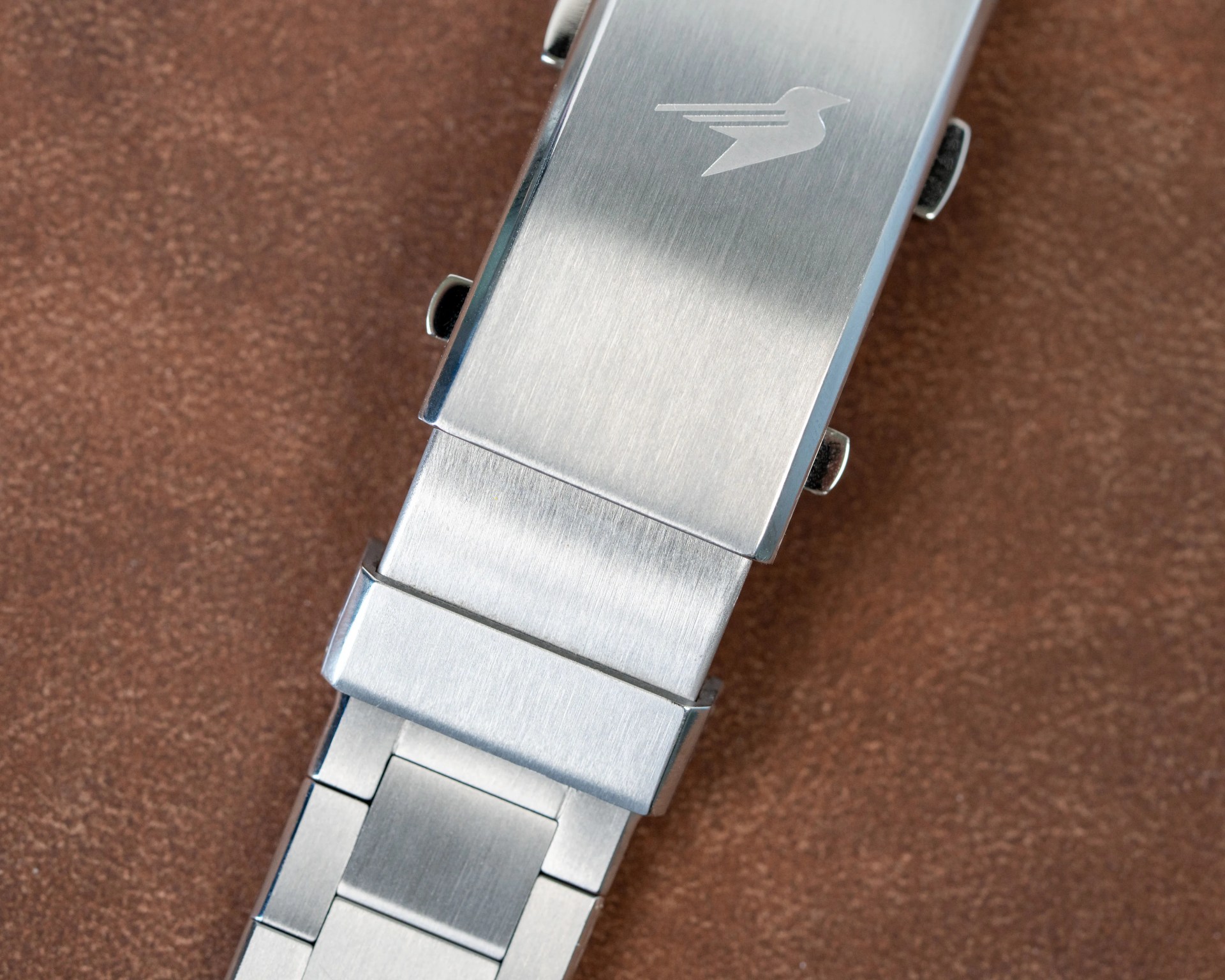 Brushed stainless steel watch clasp with an engraved bird logo on a brown textured surface.