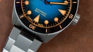 Close-up of a blue dial automatic watch with orange markers and a stainless steel bracelet on a brown surface.
