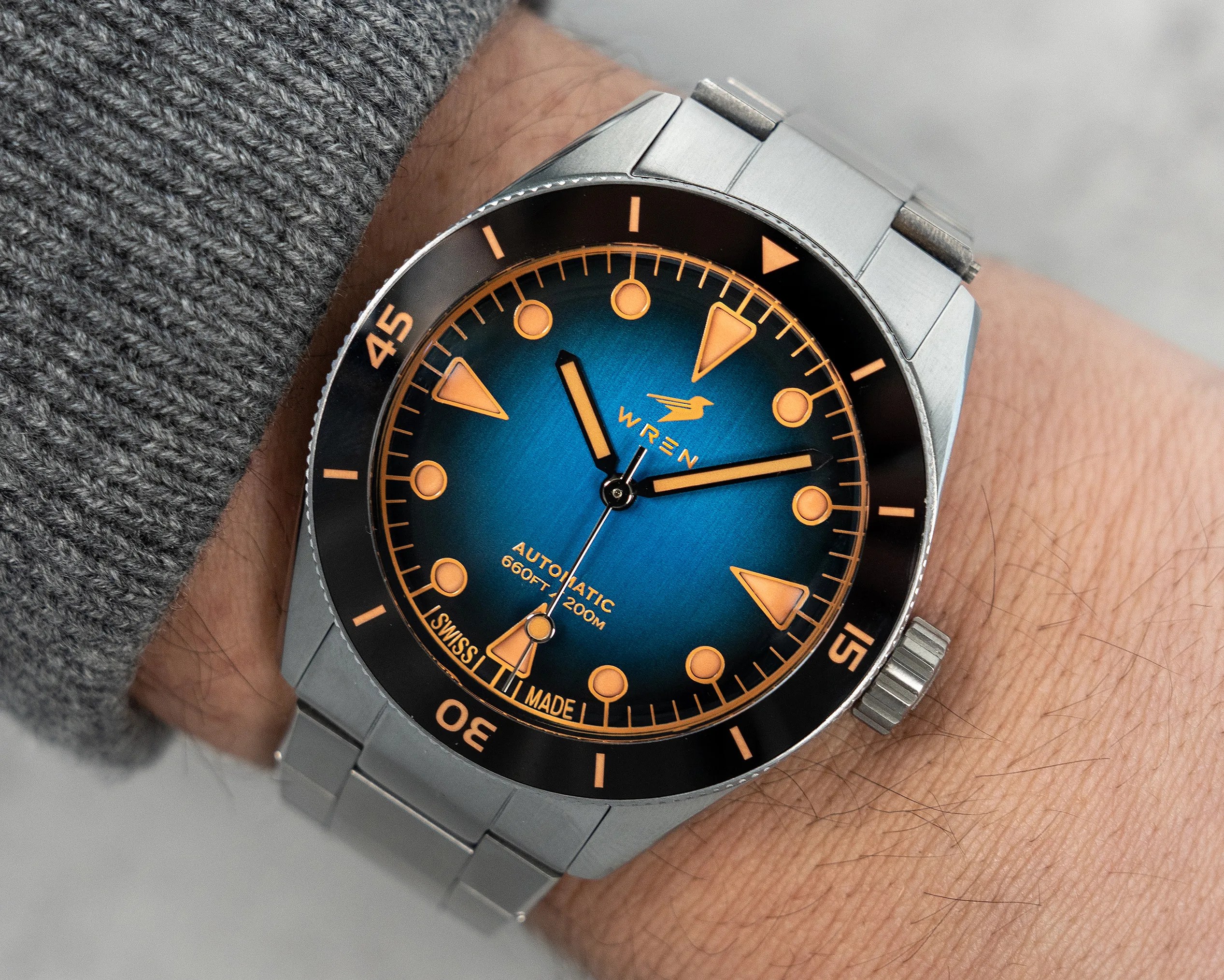 Stainless steel wristwatch with a blue dial, orange markers, black bezel, and metal bracelet on a wrist.