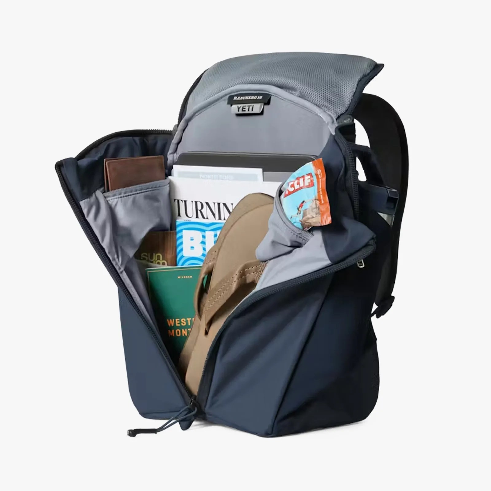 Navy blue YETI Ranchero 18 backpack open, showing organized compartments with books, wallet, flip-flop, and snack bar.