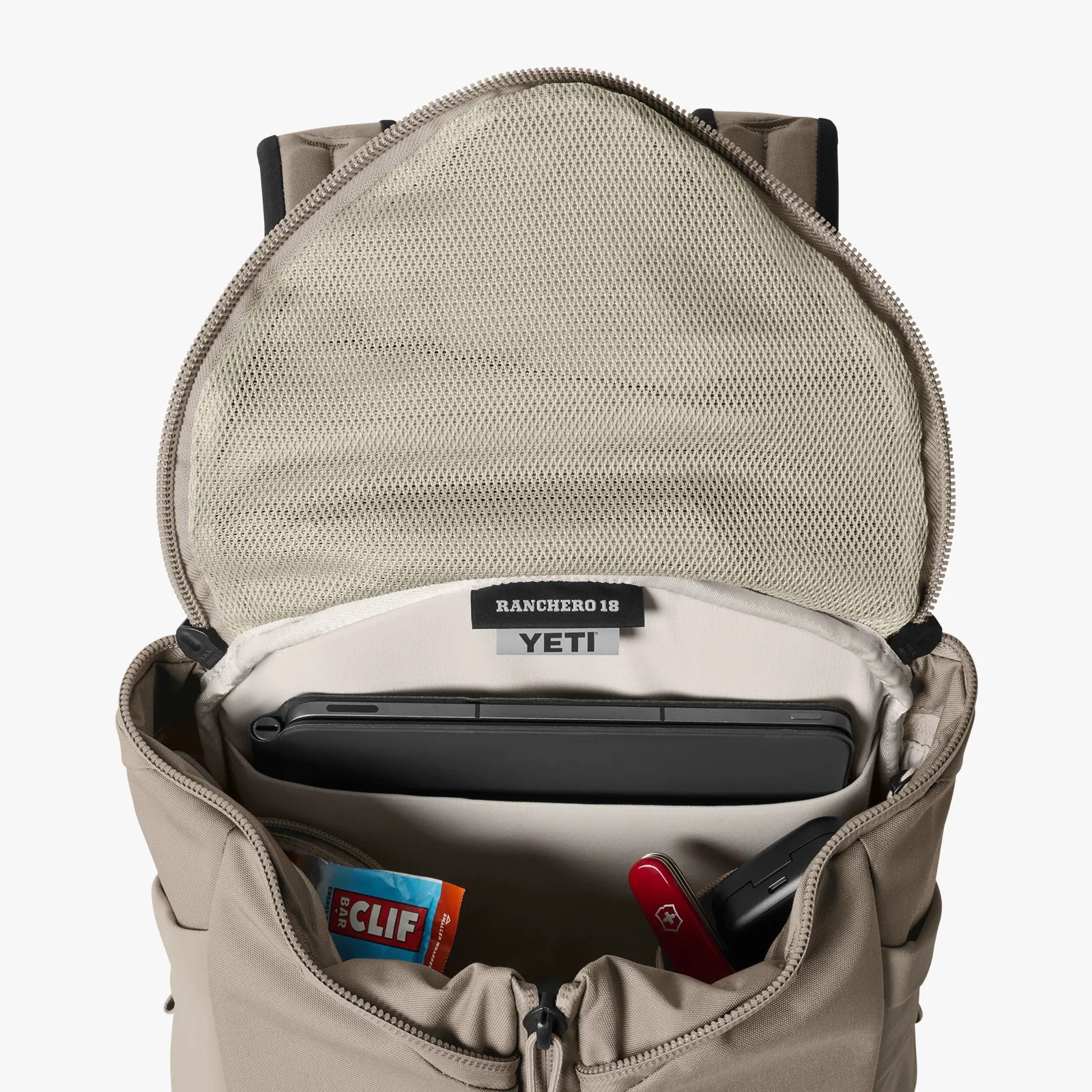 Beige YETI Ranchero 18 backpack open to show a tablet, a Clif Bar, a red Swiss Army knife, and a black case inside.