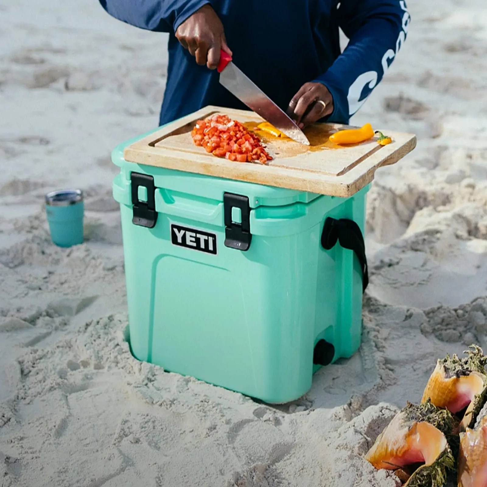 Turquoise YETI cooler on sand with wooden cutting board on top, person chopping yellow pepper and diced tomatoes.