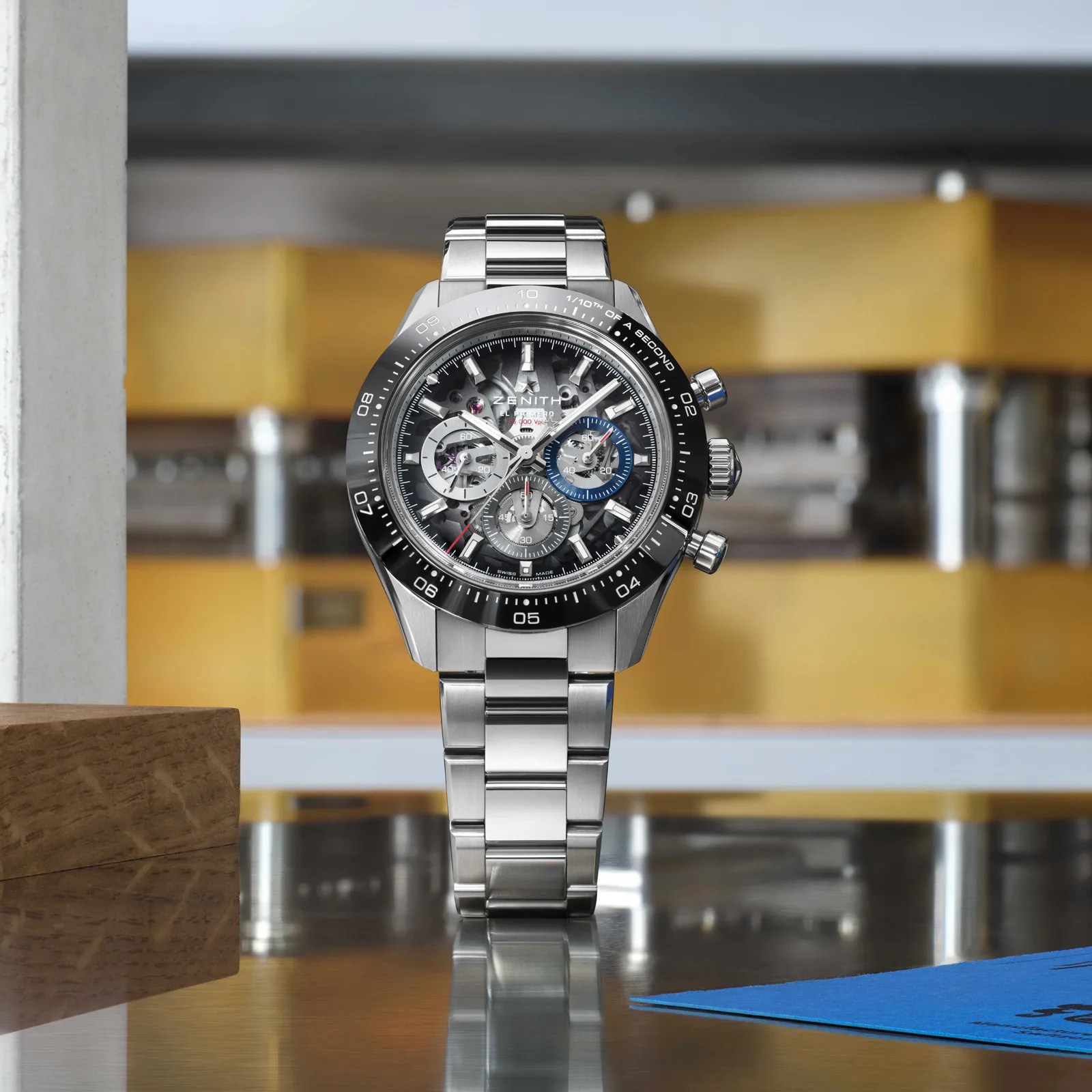Silver stainless steel Zenith chronograph watch with black bezel and skeleton dial on reflective surface.