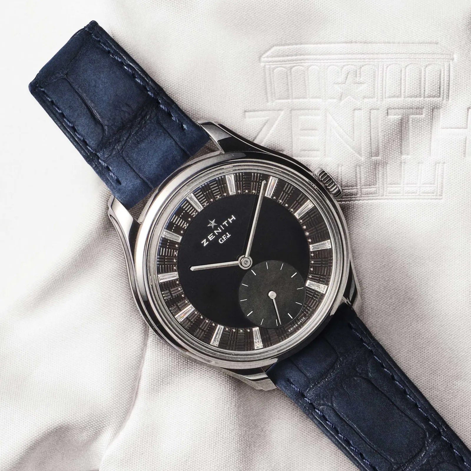 Zenith wristwatch with a black dial, silver hour markers, and a dark blue leather strap on white fabric embossed with the Zenith logo.