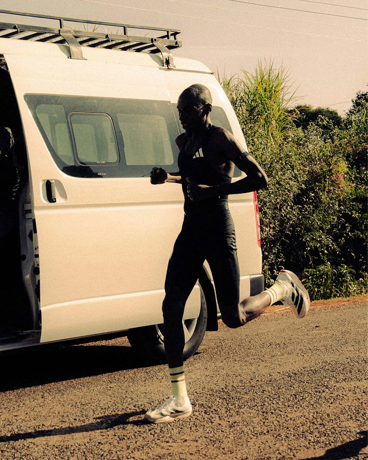 Runner in black athletic wear and white sneakers running beside a white van on a gravel road.