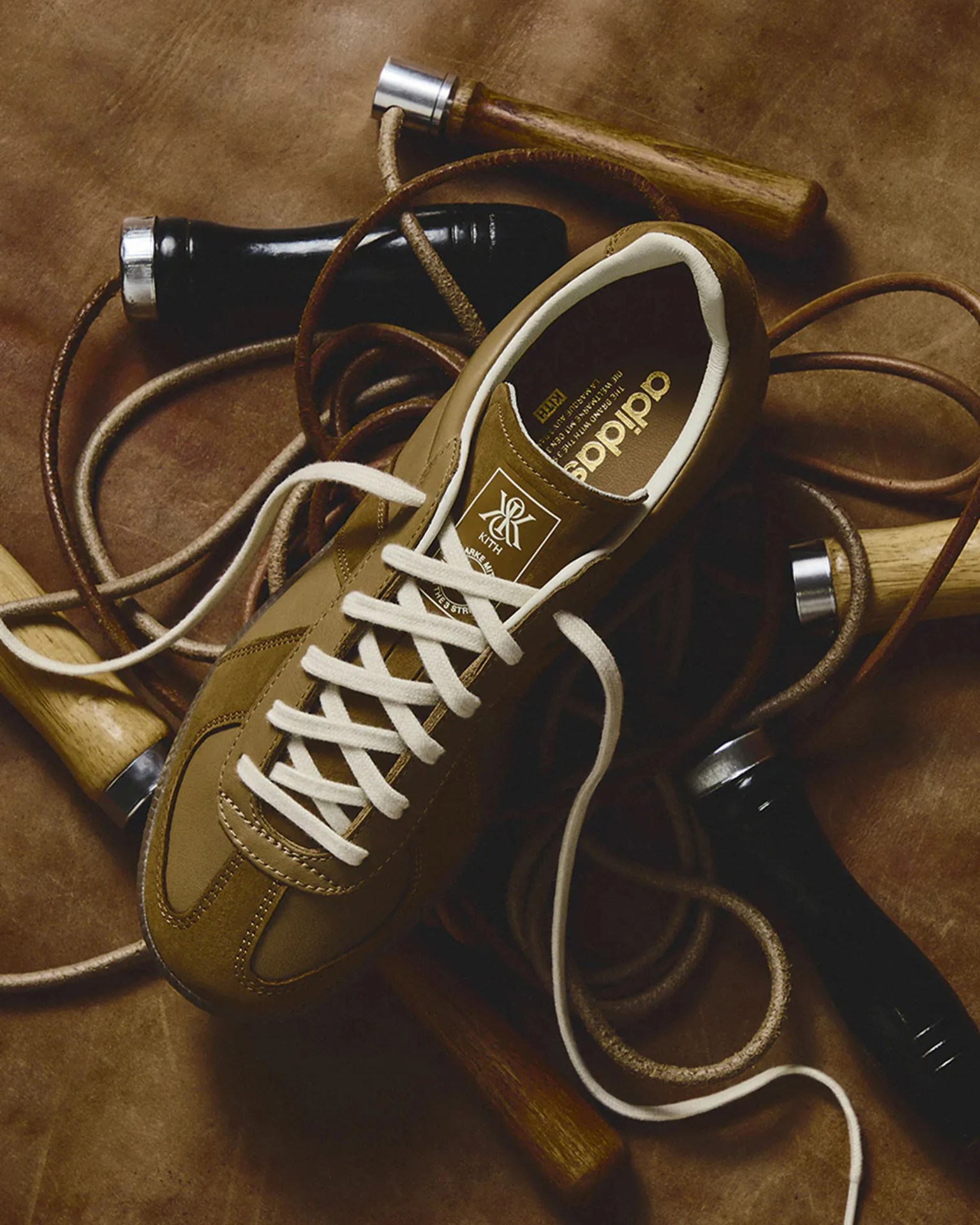 Brown Adidas sneaker with white laces and Kith logo on the tongue, placed on a brown leather surface with jump rope handles.