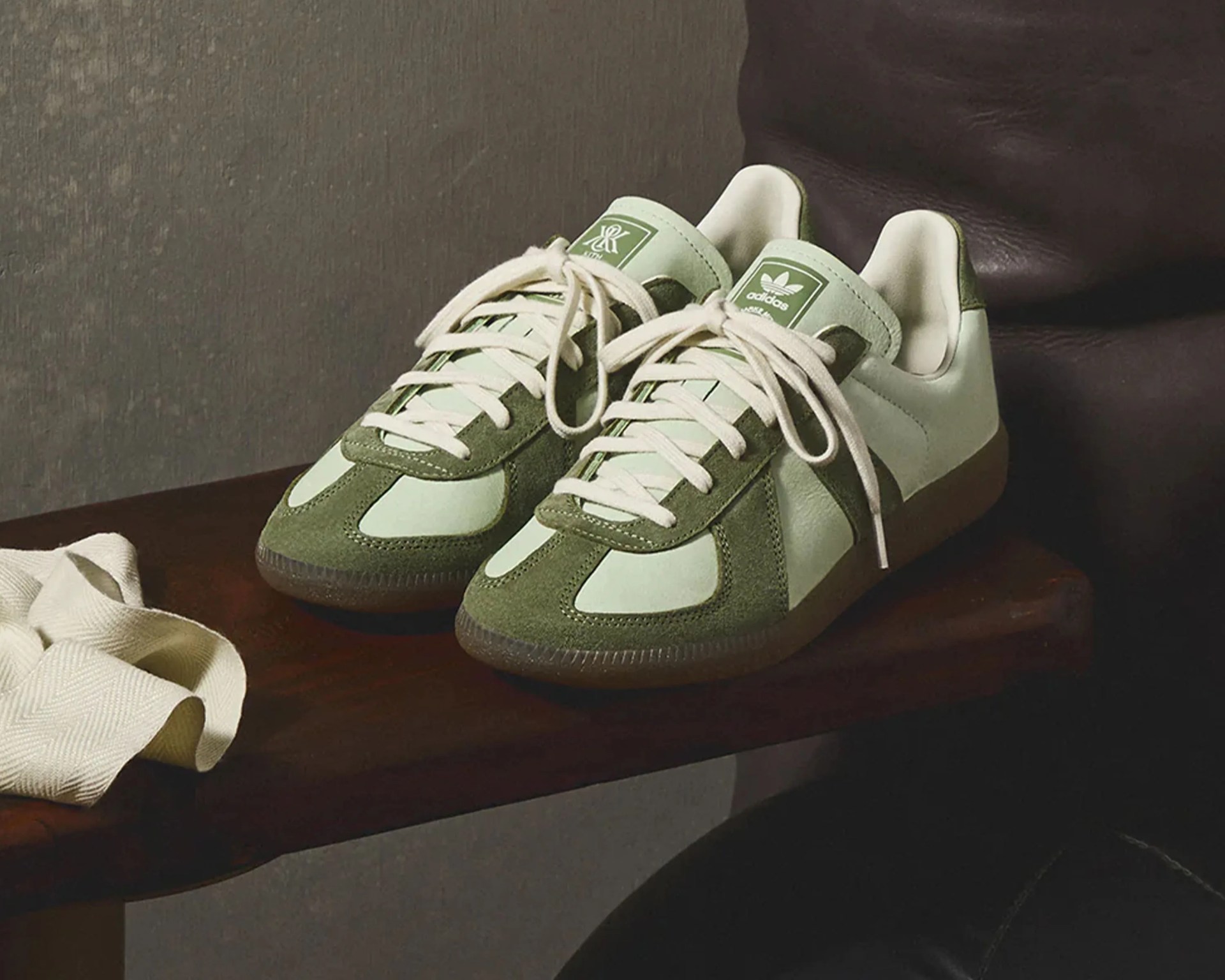 Pair of light green and olive Adidas sneakers with white laces on a wooden bench next to a cream cloth.