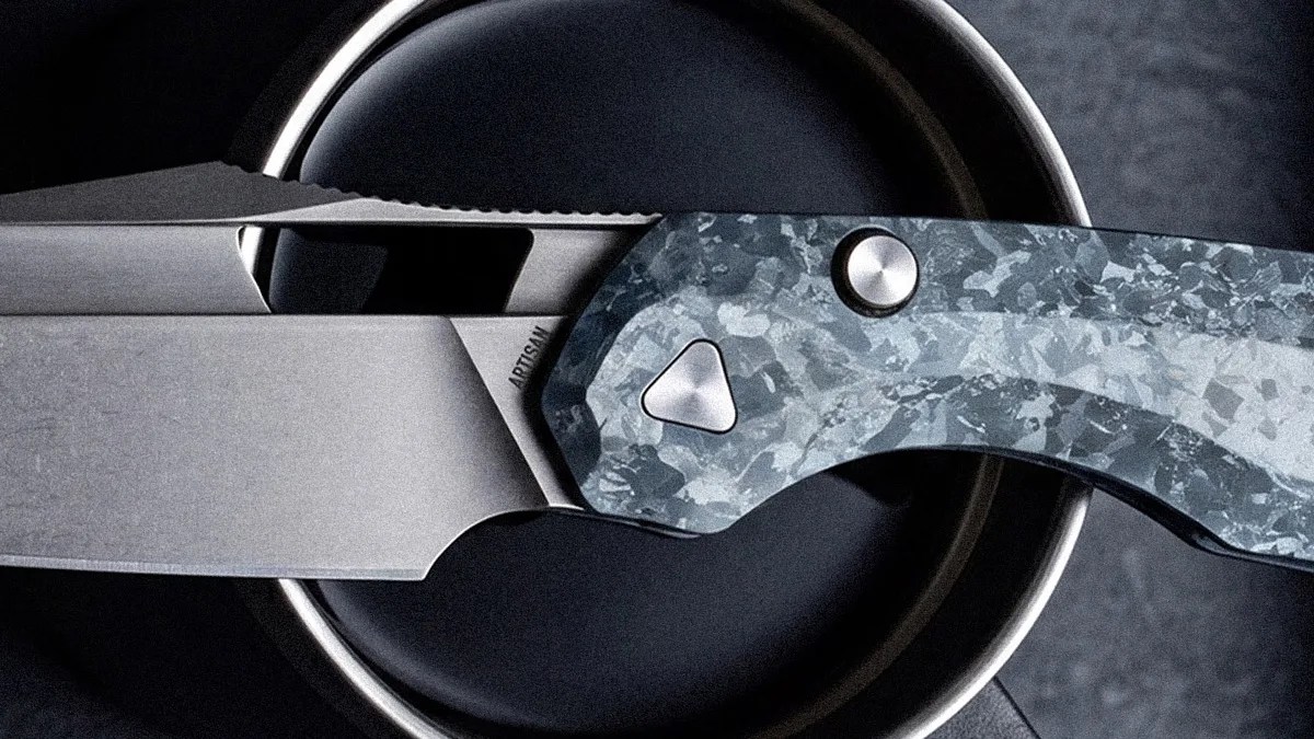 Folding knife with a stonewashed blade and a blue-gray patterned handle resting on a black circular surface.