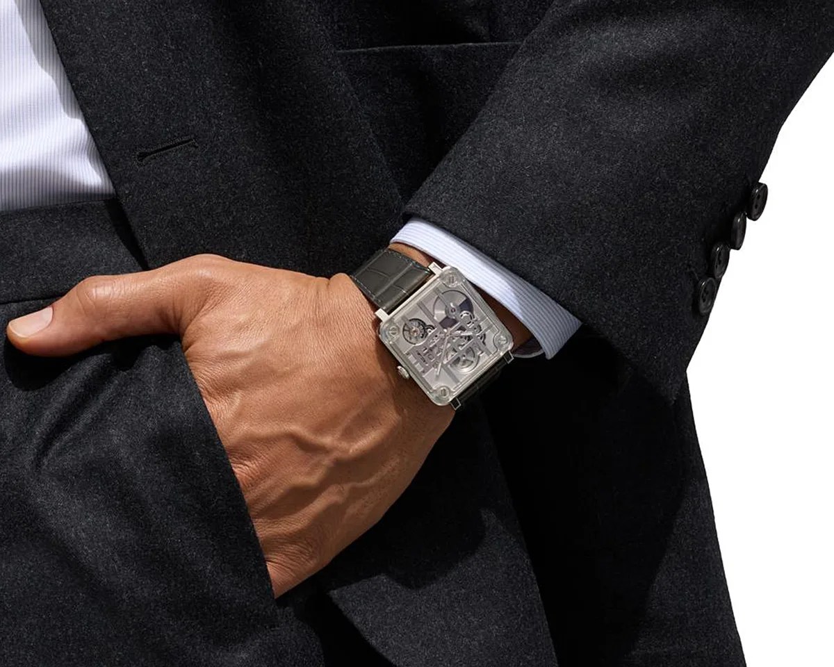 Square silver skeleton watch with black leather strap worn on wrist over white shirt cuff and dark suit sleeve.