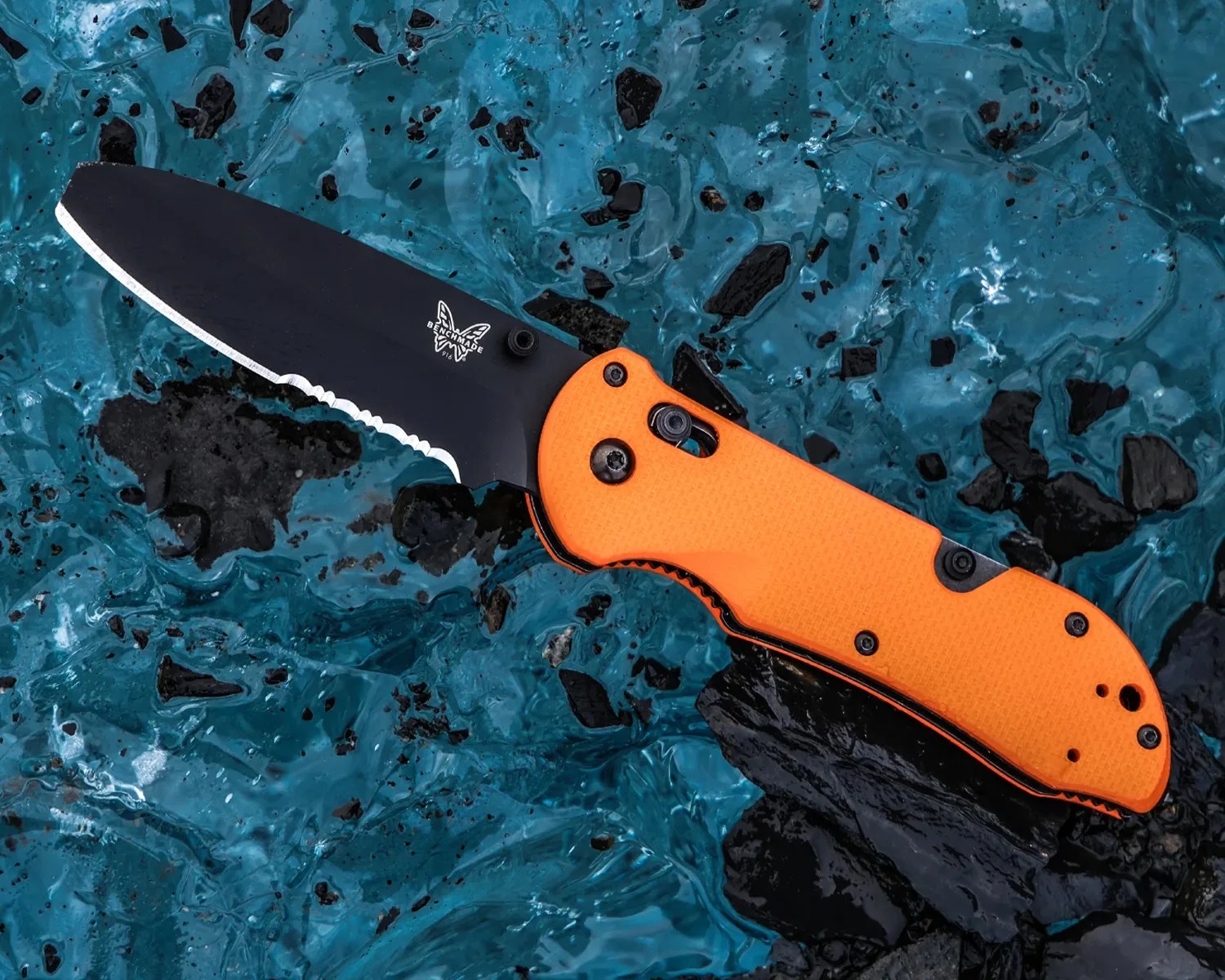 Black partially serrated Benchmade folding knife with an orange textured handle on blue and black rocky surface.