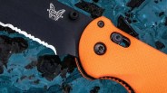 Black partially serrated Benchmade 916 knife blade with orange textured handle on a blue and black surface.