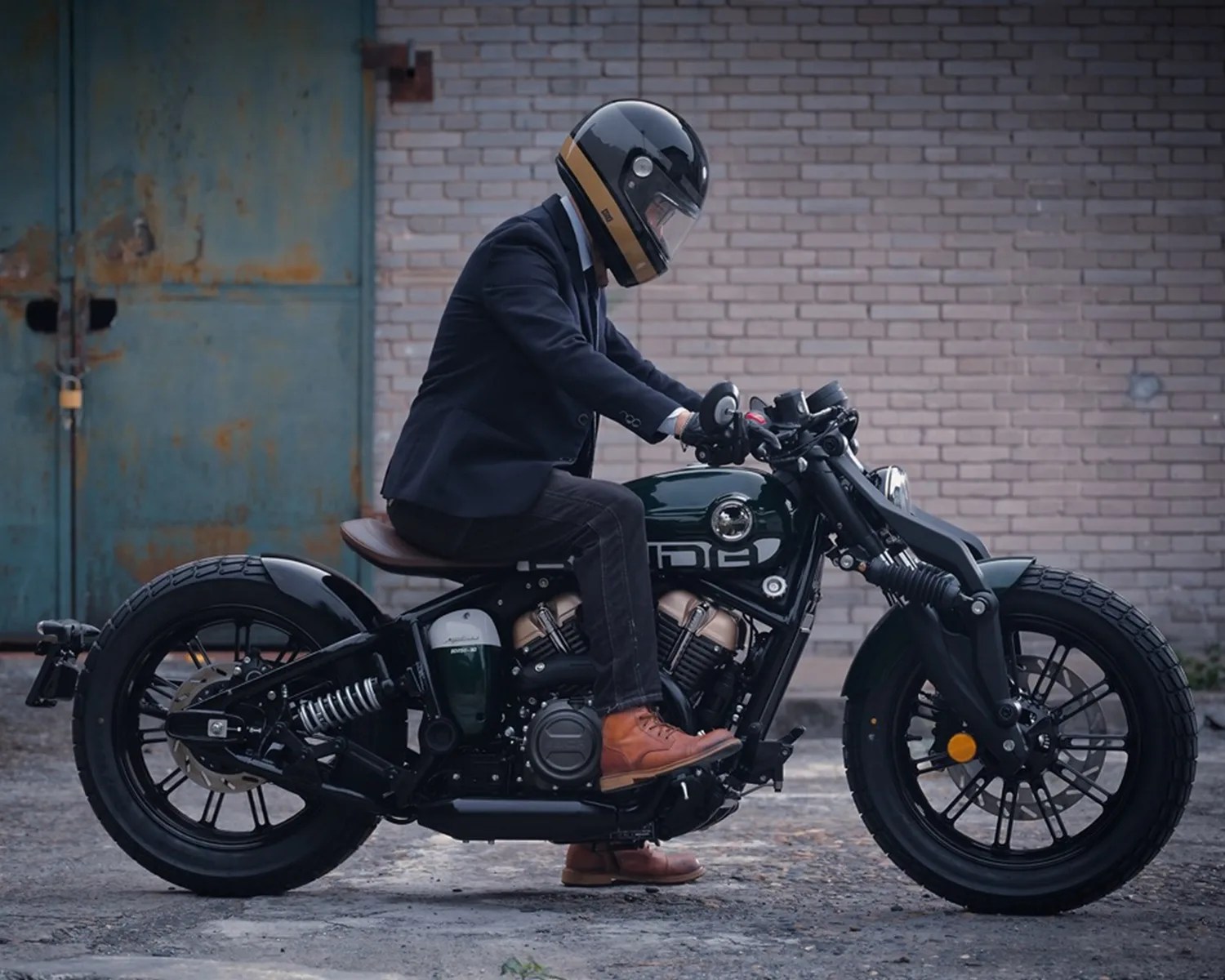 Person wearing a black and gold helmet, navy blazer, and brown boots sitting on a black motorcycle with a brown leather seat.
