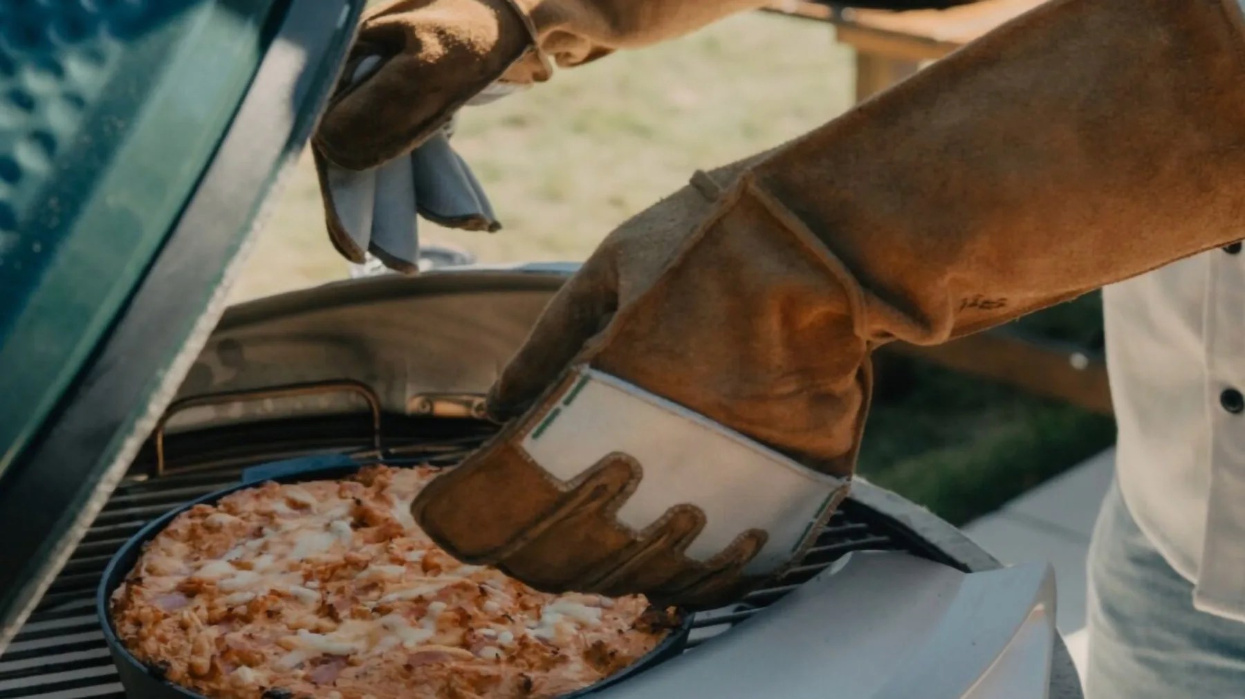 Person wearing brown leather gloves removing a cooked pizza from a green outdoor grill.