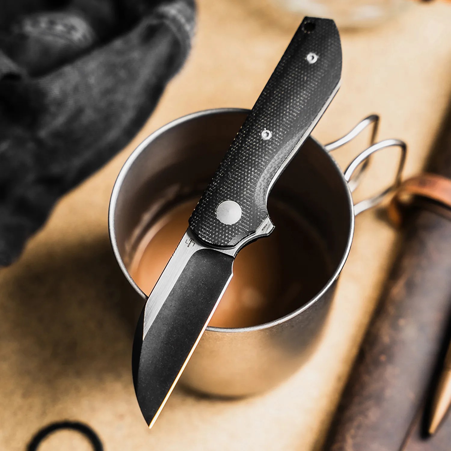 Folding knife with black textured handle resting on a metal cup.