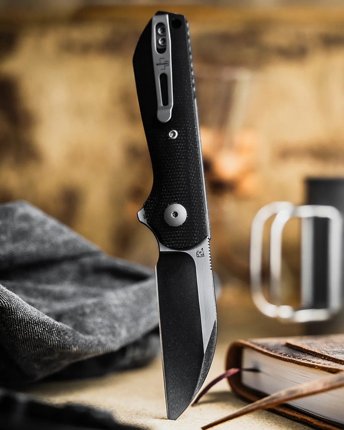 Black folding knife with textured handle standing upright on a surface next to a closed book and fabric.