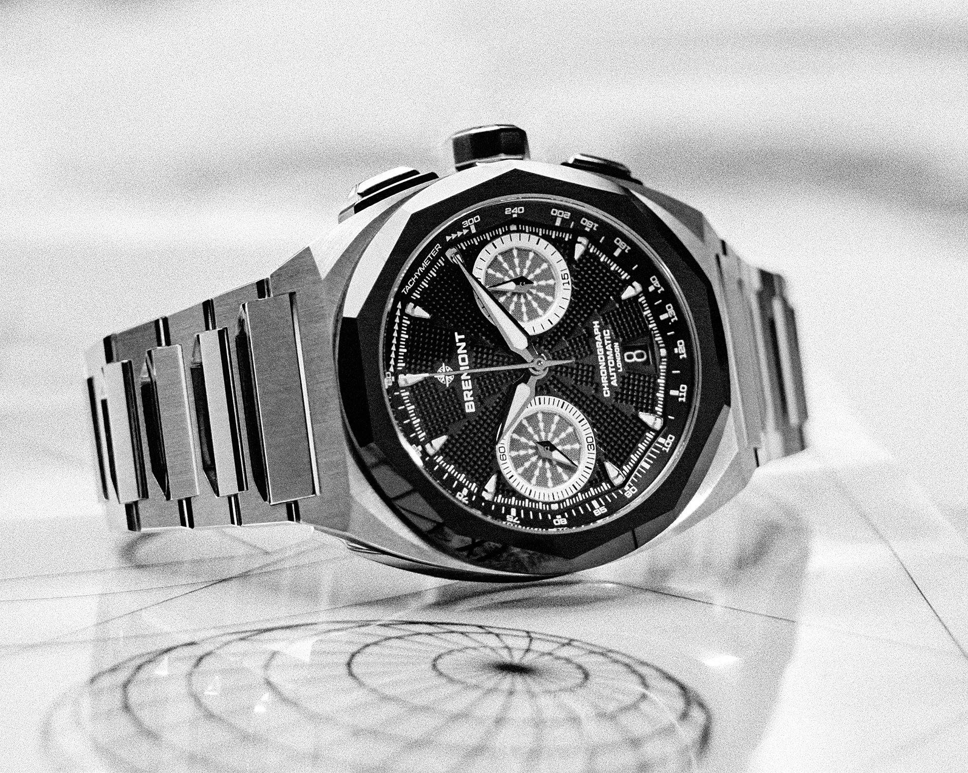 Silver metal chronograph wristwatch with black dial and two subdials resting on a reflective surface.