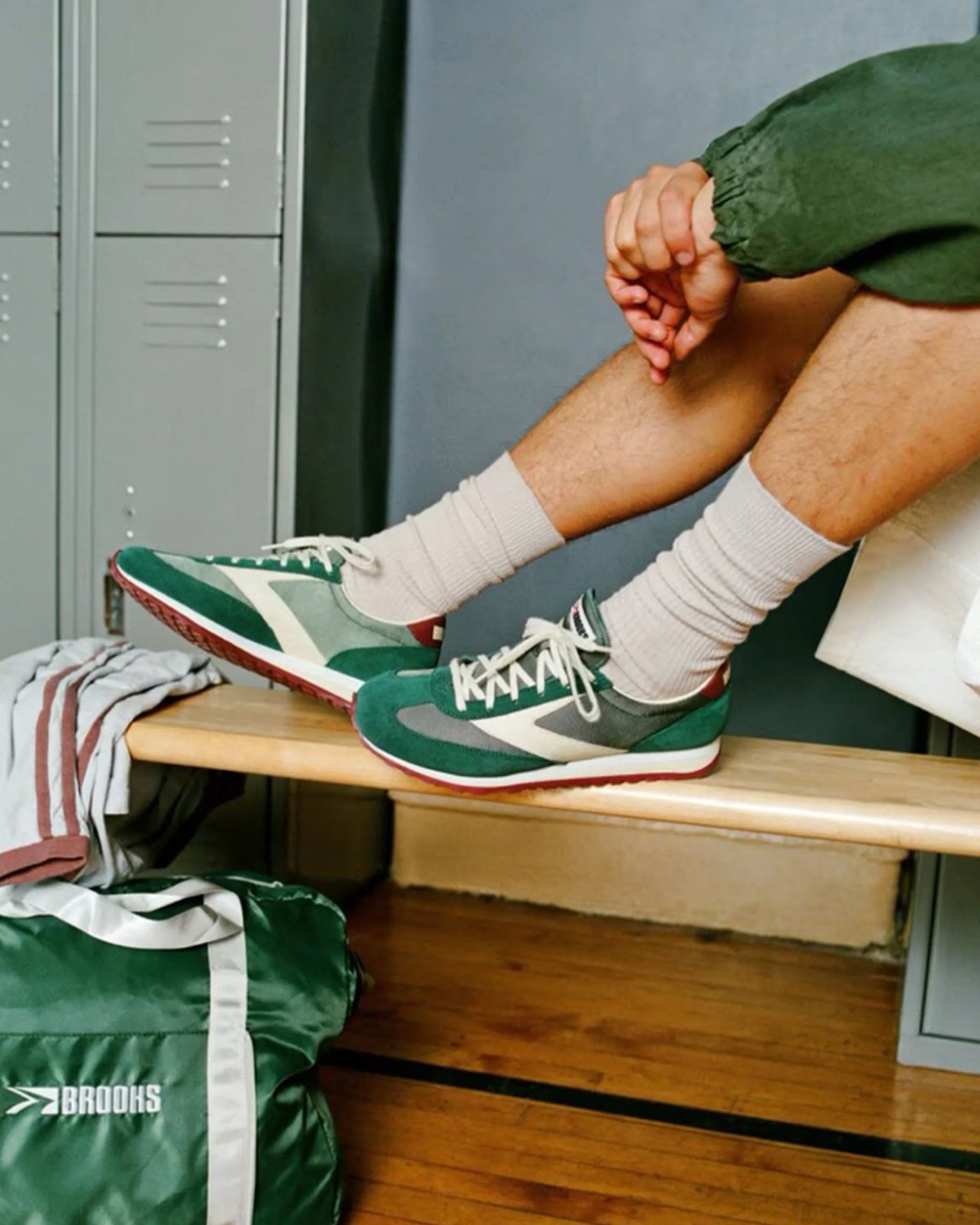 Green and gray Brooks running shoes with white laces worn with beige socks, shown on a wooden bench in a locker room.