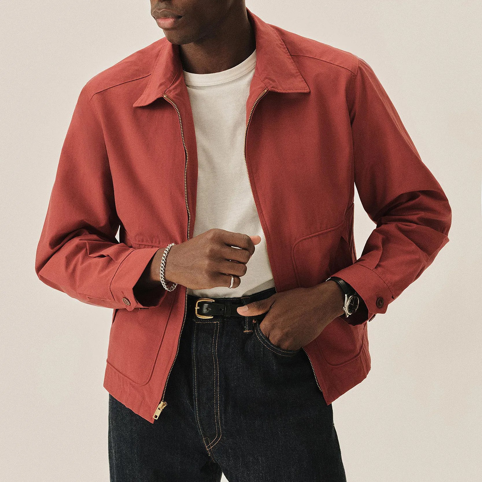 Rust-colored zip-up jacket worn over a white t-shirt with dark denim jeans and black belt.