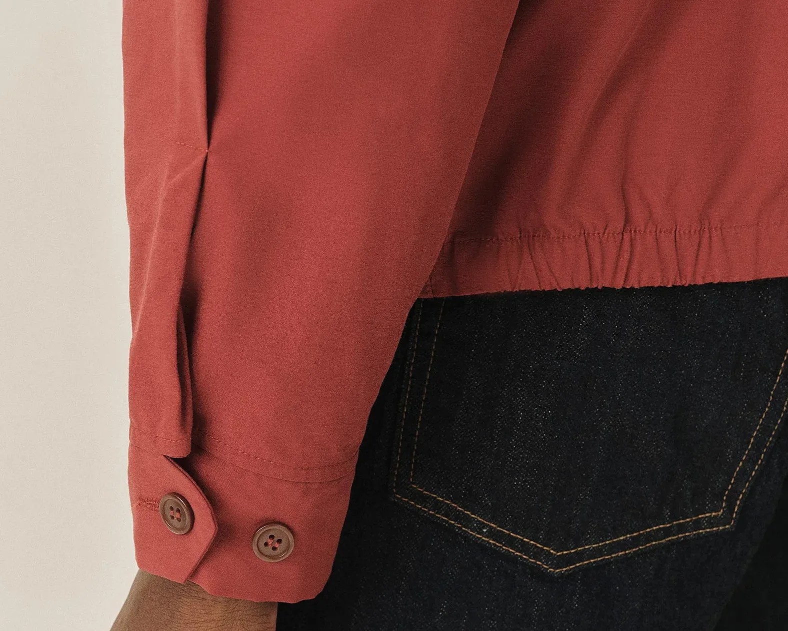 Rust-colored long sleeve shirt cuff with two brown buttons worn over dark blue denim jeans pocket.