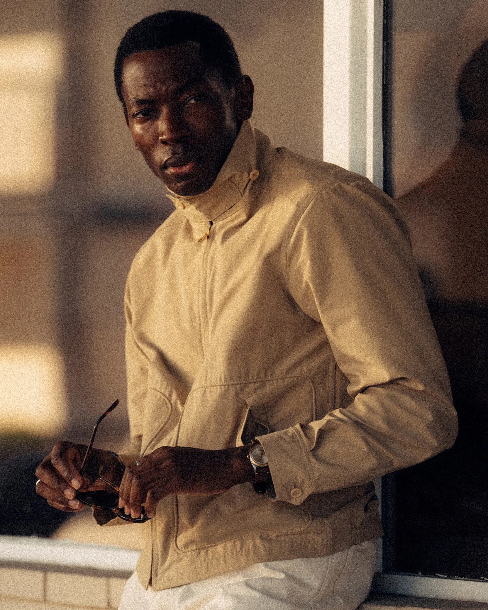 Man wearing a beige zip-up jacket holding sunglasses near a window with a watch on his wrist.