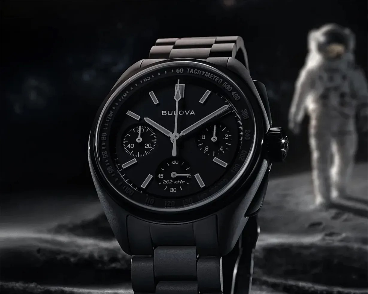 Black Bulova chronograph watch with tachymeter bezel and metal link band, astronaut figure blurred in background.
