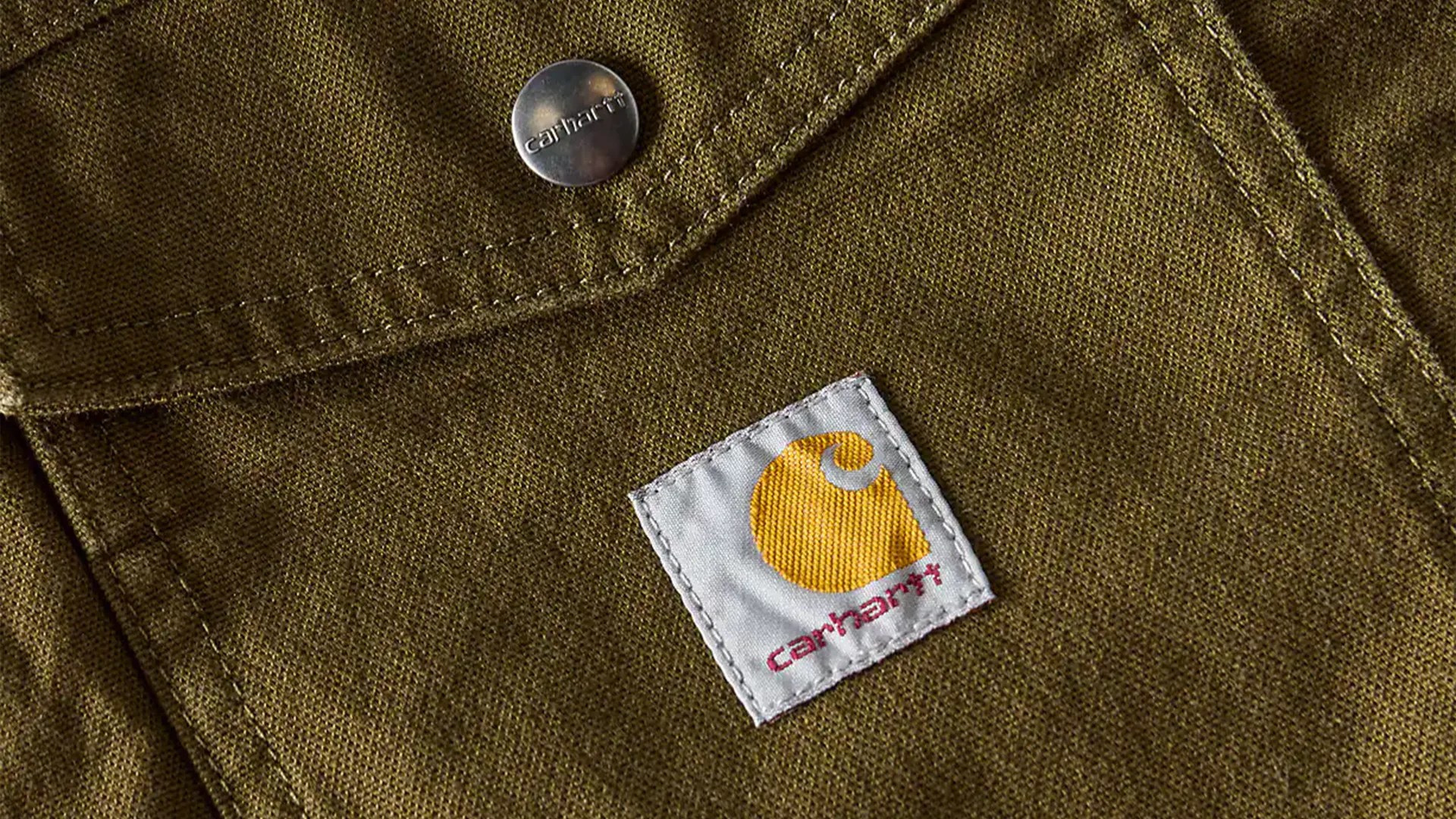 Close-up of olive green Carhartt fabric with a white and yellow Carhartt logo patch and a metal button embossed with "carhartt".
