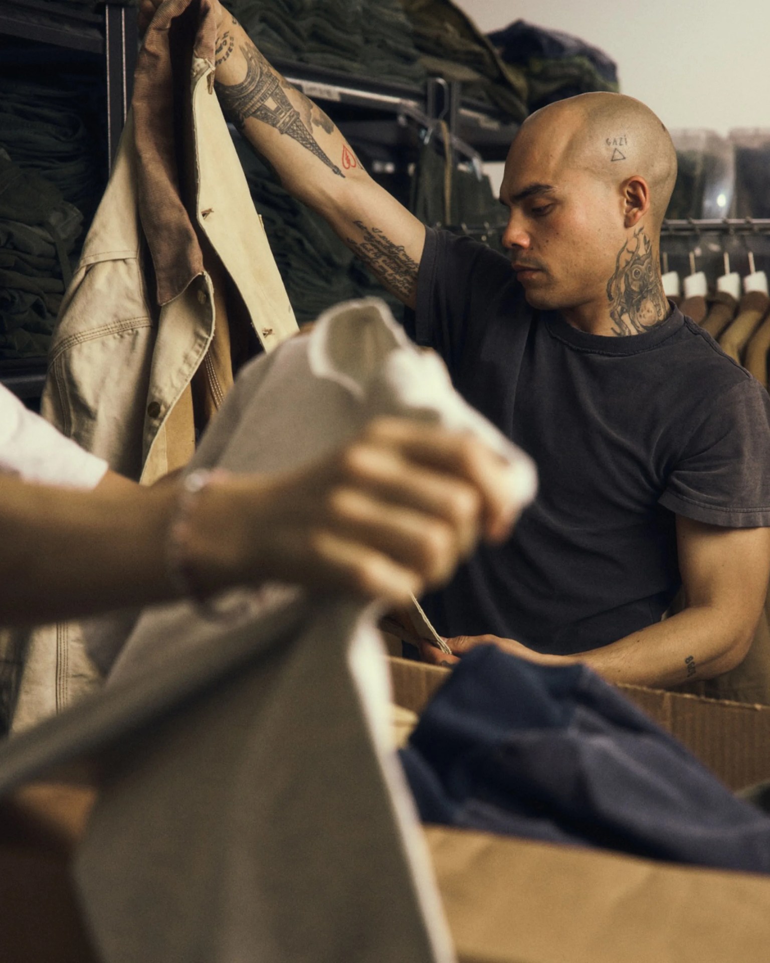 Person with tattoos on head and arms holding a beige jacket in a clothing store.