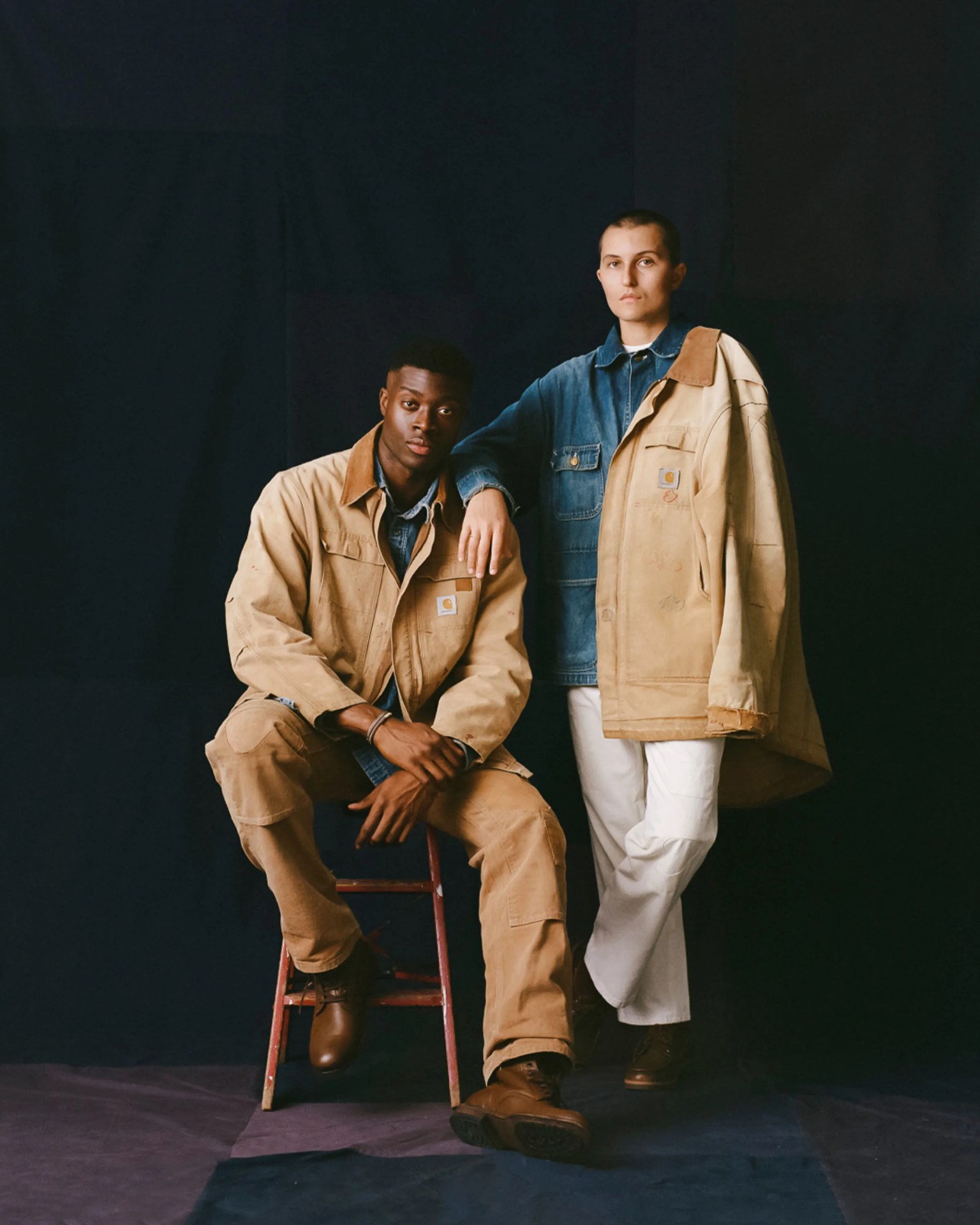 Two models wearing tan Carhartt jackets and brown boots, one seated on a stool and the other standing with white pants against a dark backdrop.
