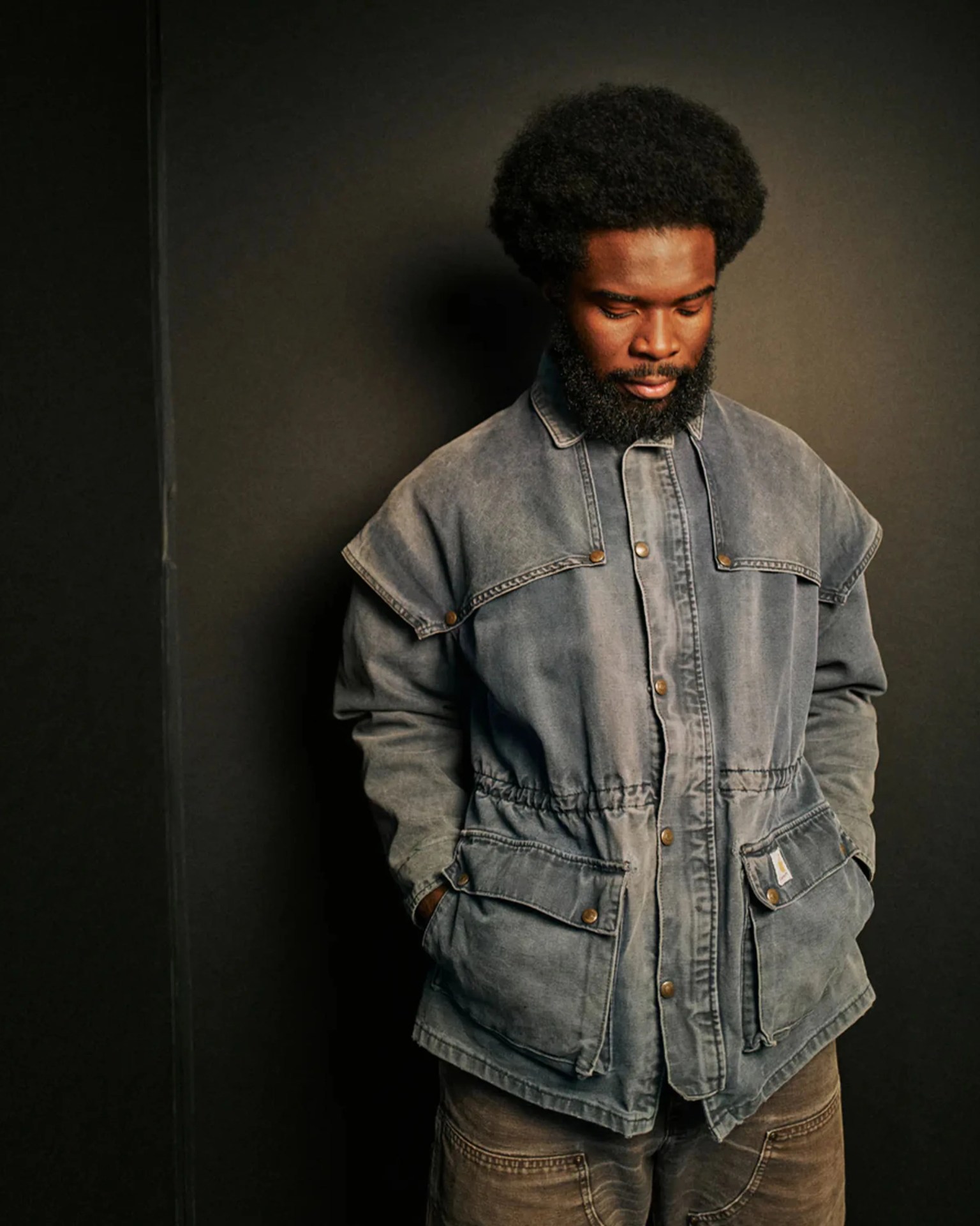 Man wearing a faded blue denim jacket with snap buttons and large front pockets against a dark background.