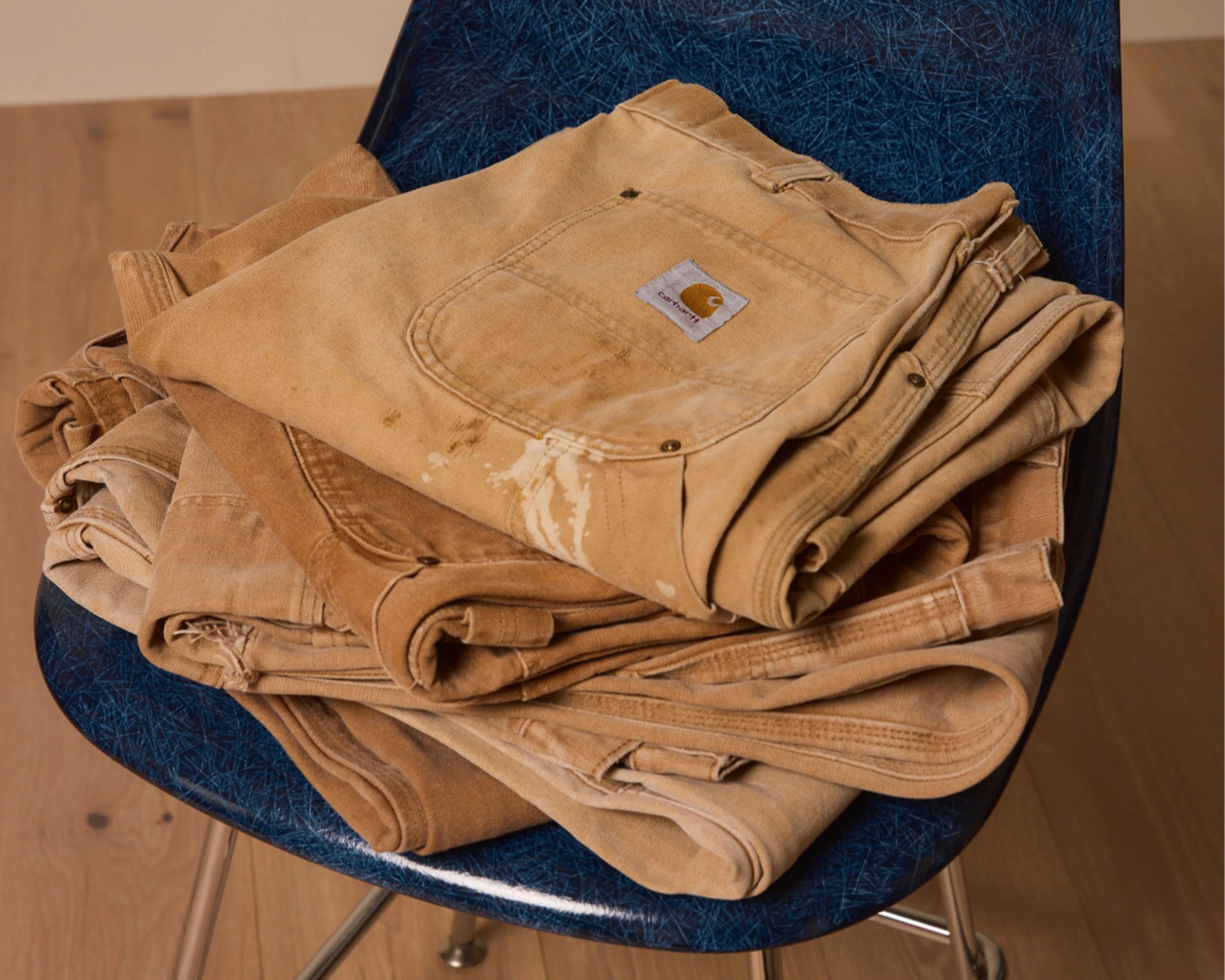Stack of folded tan and brown Carhartt work pants on a blue chair with metal legs.