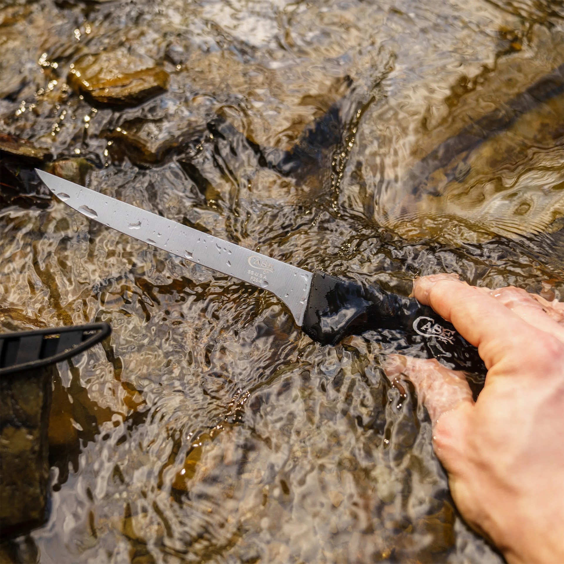 Hand holding a black-handled Case brand knife with a wet blade submerged in shallow water over rocks.