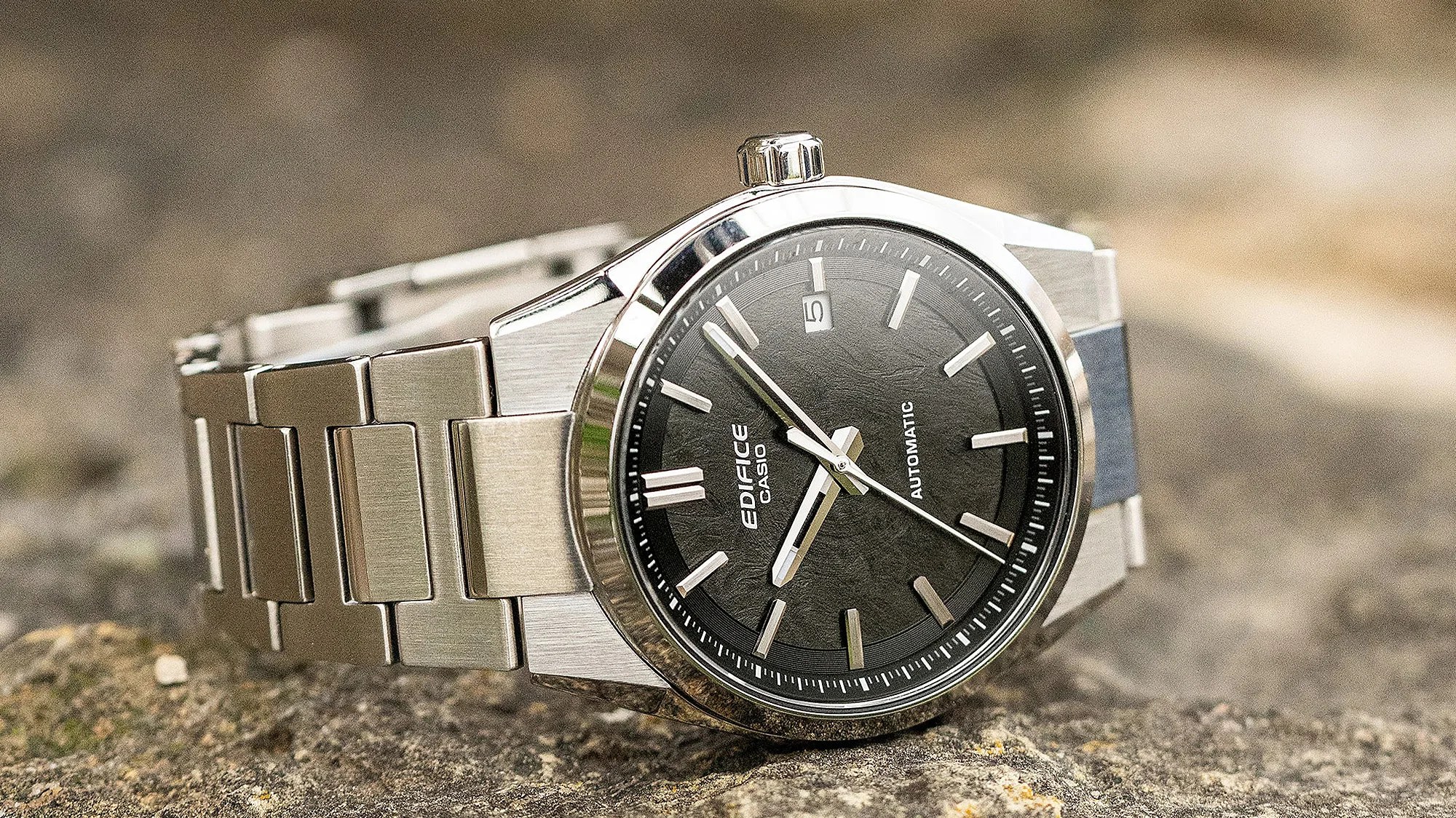 Silver Casio Edifice automatic wristwatch with black textured dial and metal link bracelet on a stone surface.