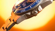 Close-up of a stainless steel wristwatch with a blue bezel and crown engraved with "GS" against an orange background.