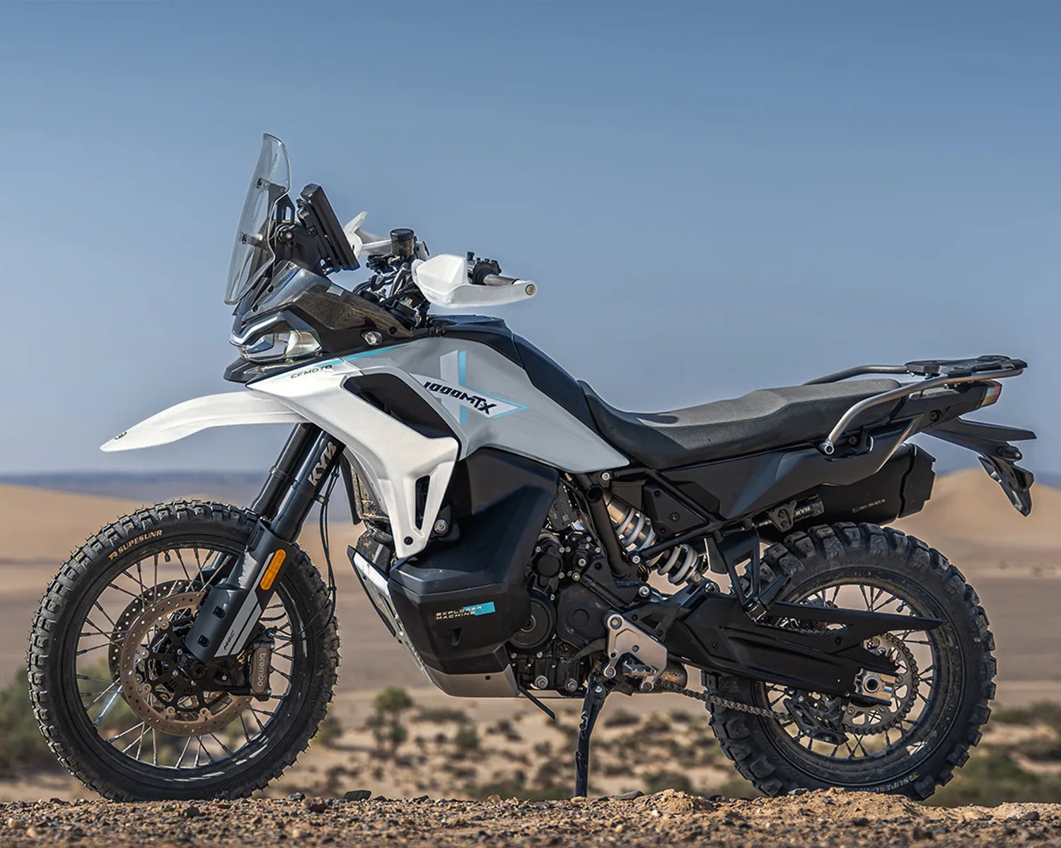 White and black CFMOTO 1000MT-X adventure motorcycle with knobby tires parked on rocky terrain.