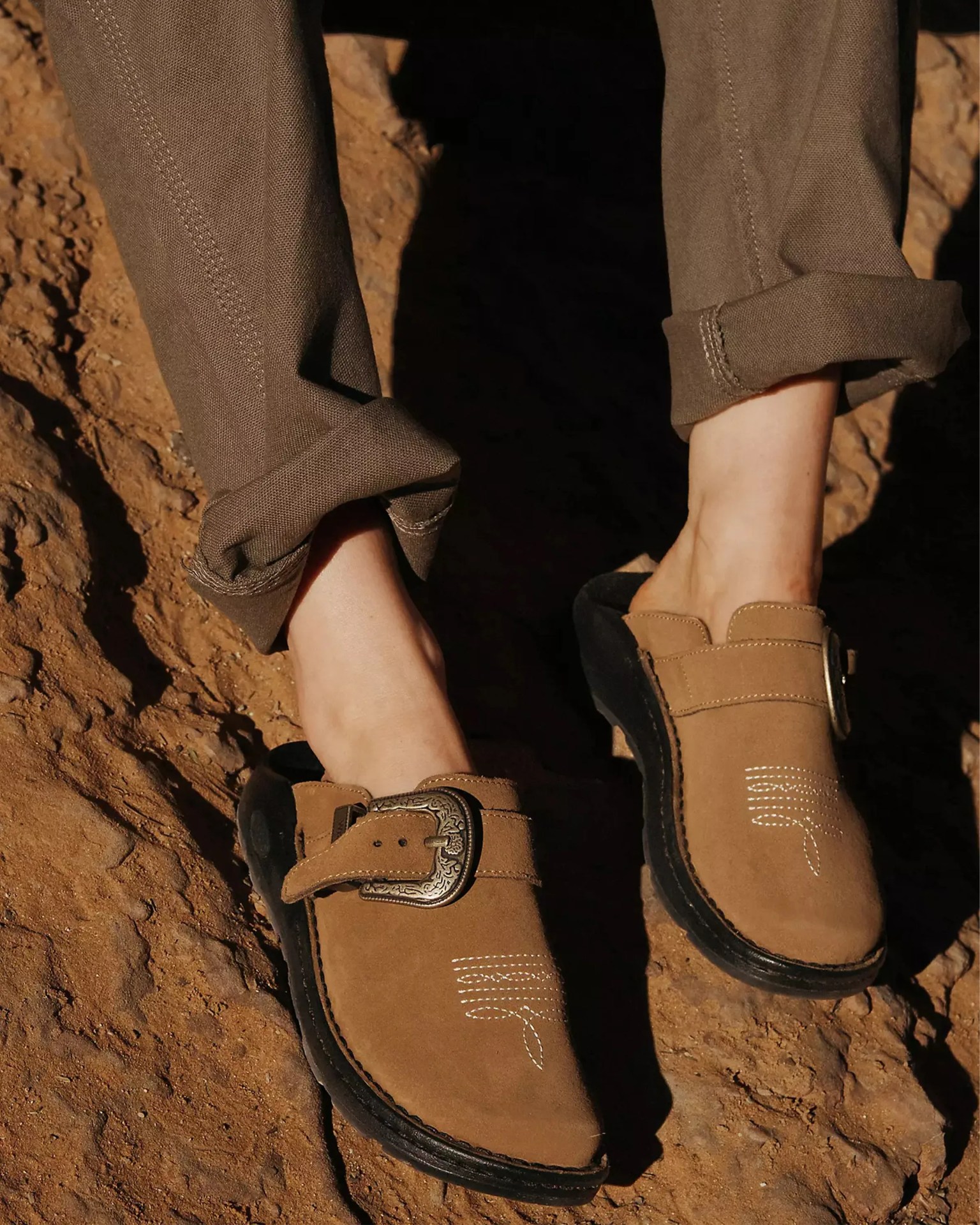 Brown suede slip-on shoes with decorative stitching and metal buckle on a dirt ground.