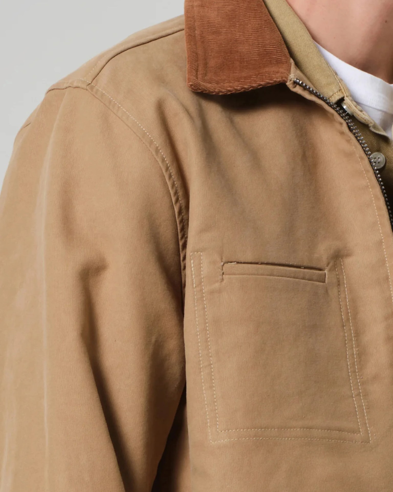 Tan canvas jacket with brown corduroy collar and front chest pocket, partially zipped over a white shirt.