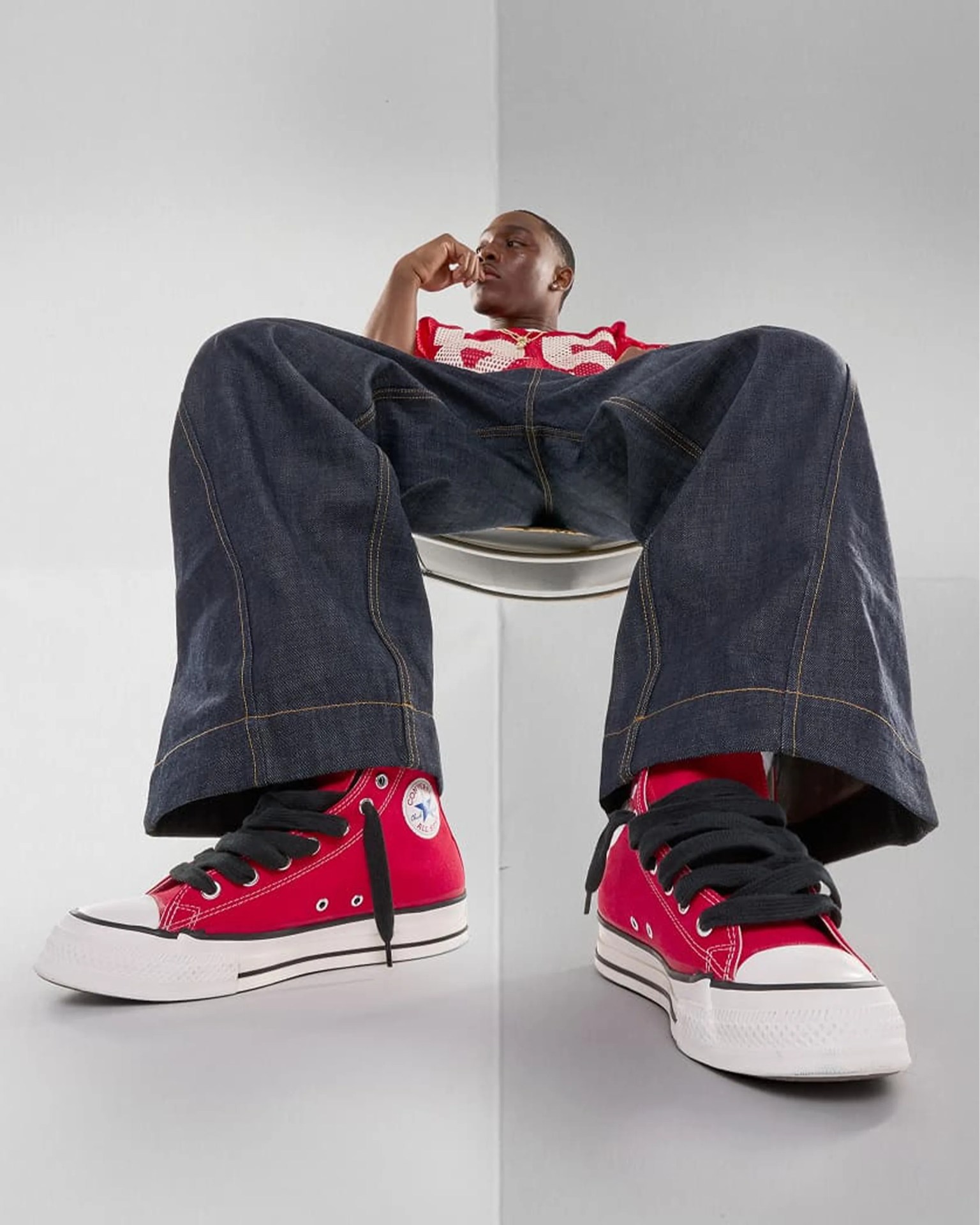 Person wearing dark blue wide-leg jeans and red Converse high-top sneakers with black laces sitting on a chair.