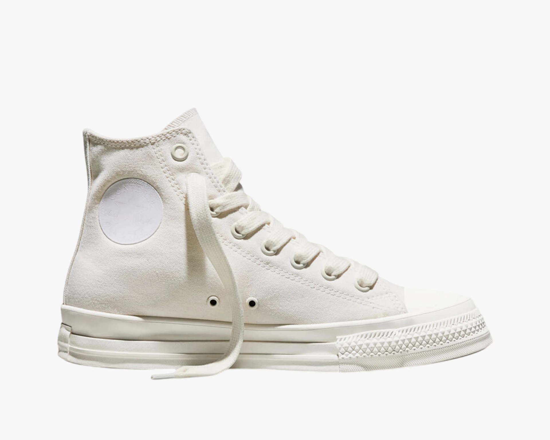 Off-white high-top canvas sneaker with white rubber sole and laces, featuring a subtle circular patch on the ankle.