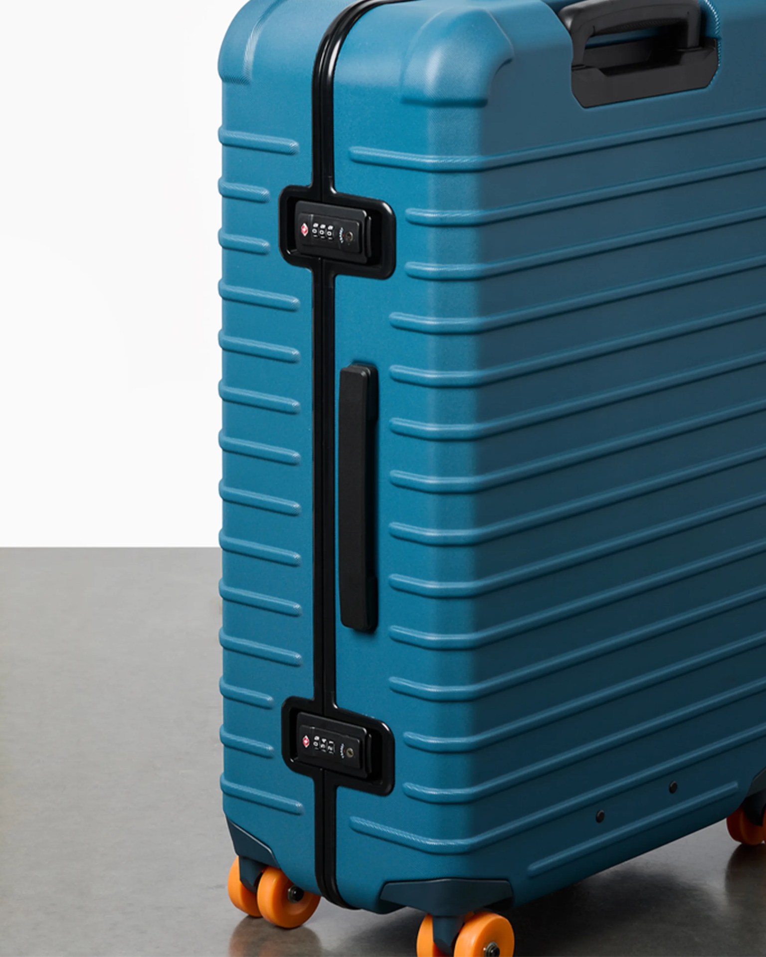 Blue hard-shell suitcase with black handle, black zipper, built-in combination locks, and orange wheels on a gray floor.