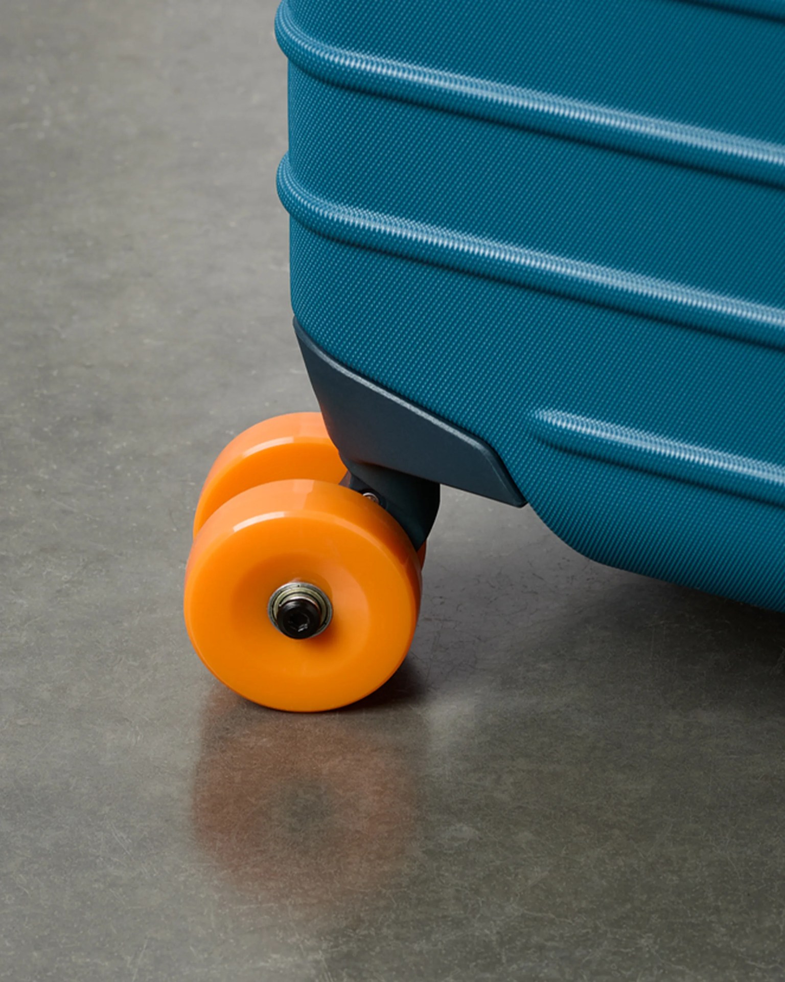 Close-up of a teal hard-shell suitcase corner with bright orange double wheels on a gray floor.
