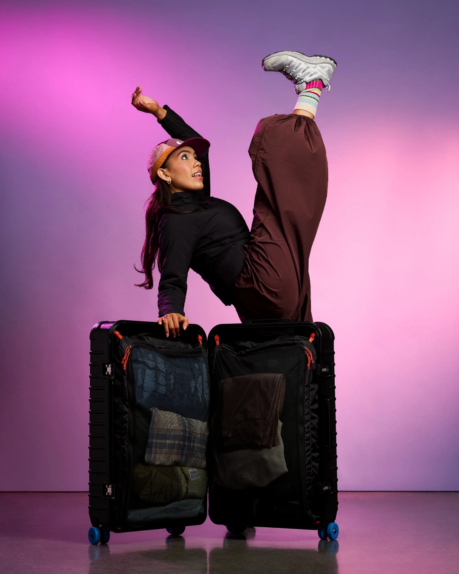 Person wearing a black jacket, brown pants, and colorful socks with gray sneakers balancing on an open black suitcase with blue wheels.
