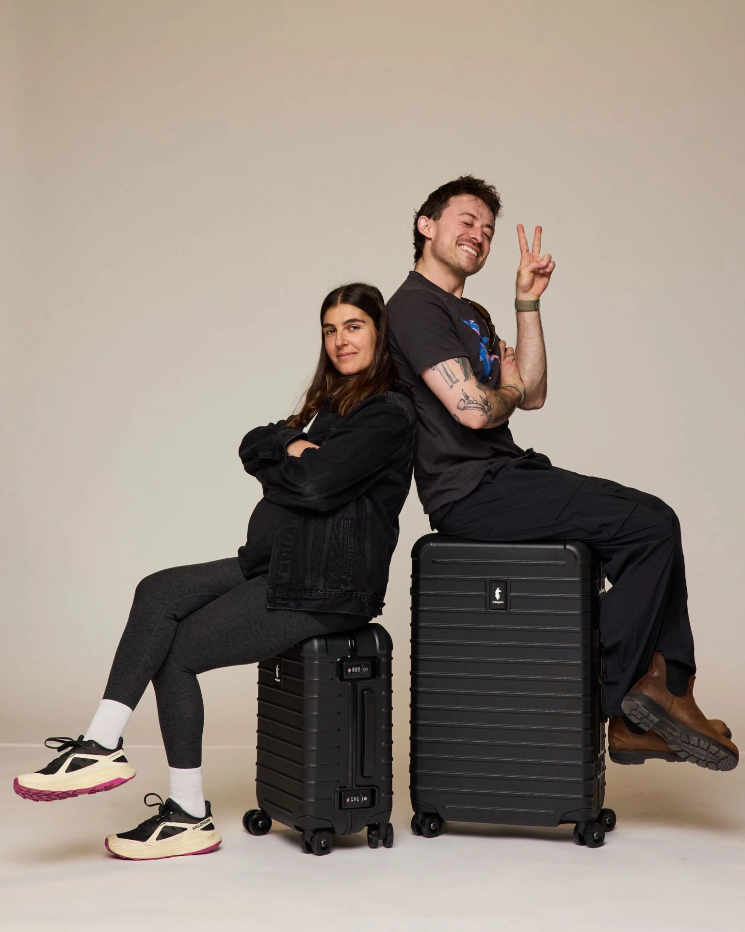 Two people sitting back-to-back on black hard-shell suitcases, one making a peace sign.