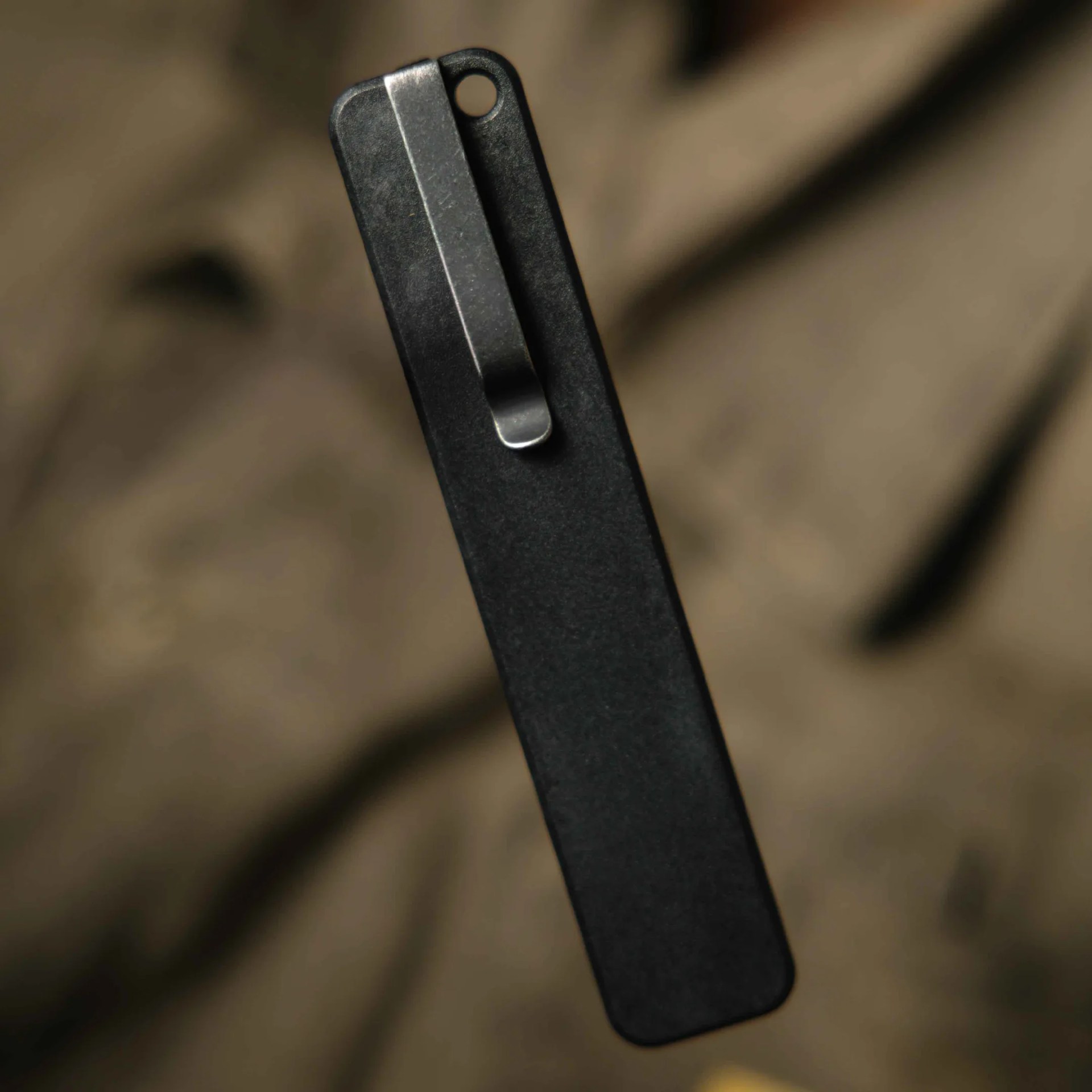 Black rectangular metal clip with a silver spring clip attached on a blurred brown background.