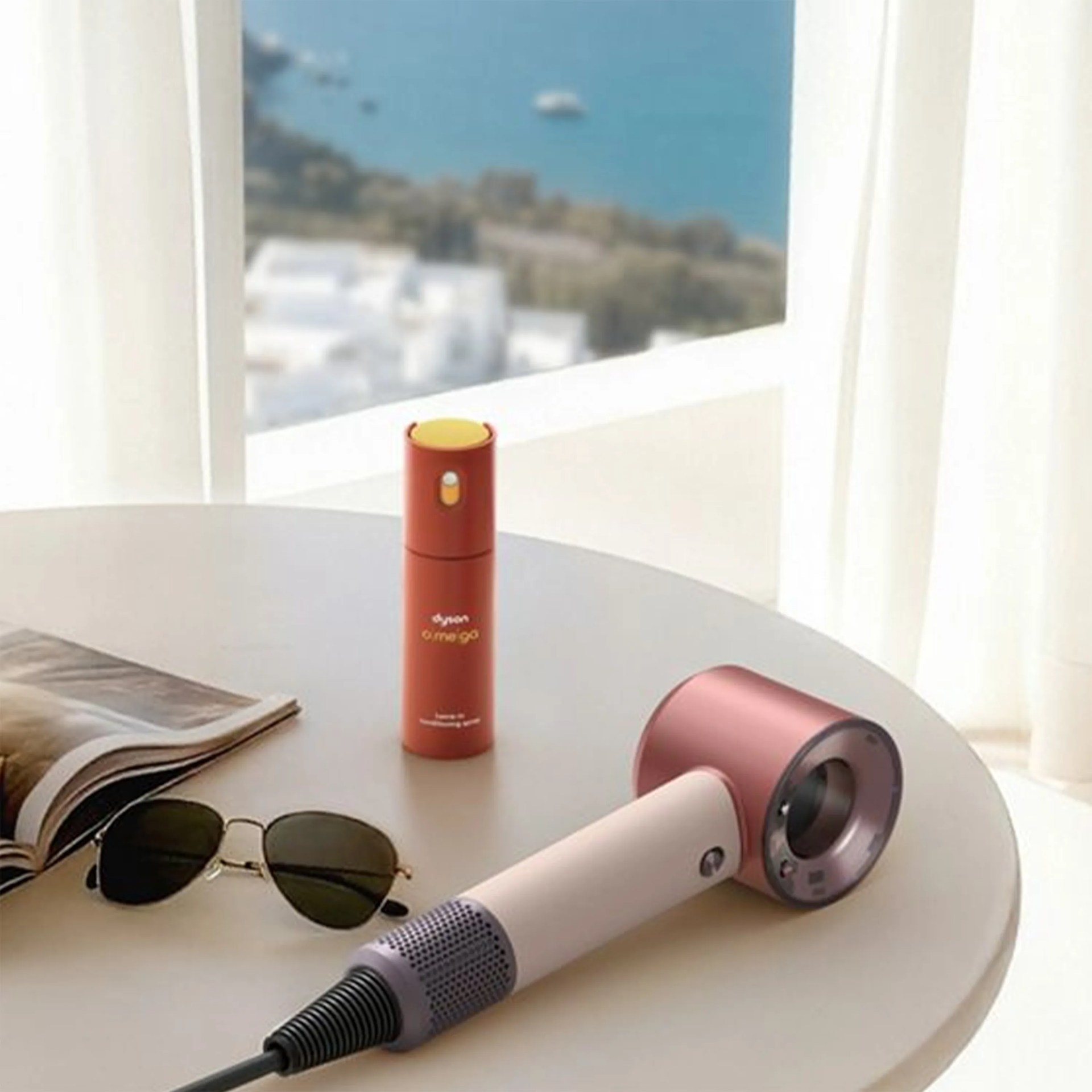 White and pink Dyson hair dryer, red Dyson Omigo fragrance bottle, sunglasses, and open magazine on a round table.