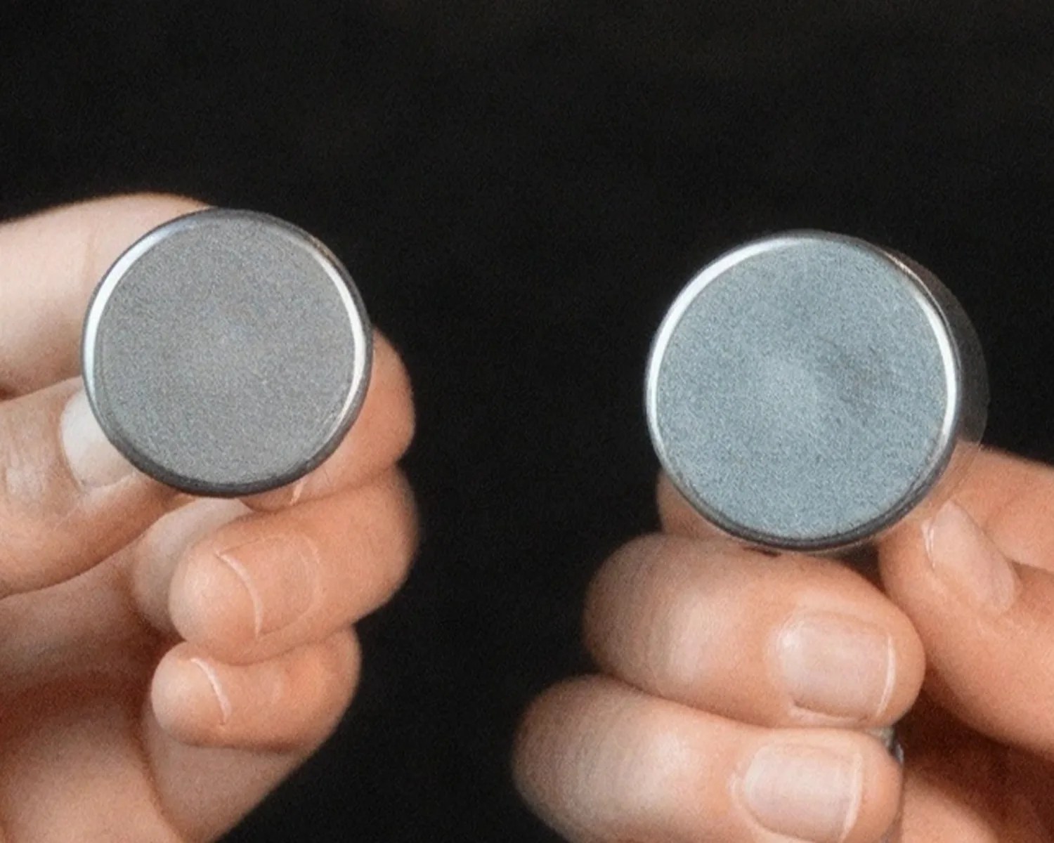 Two hands holding round, silver-colored metal objects with flat tops against a black background.